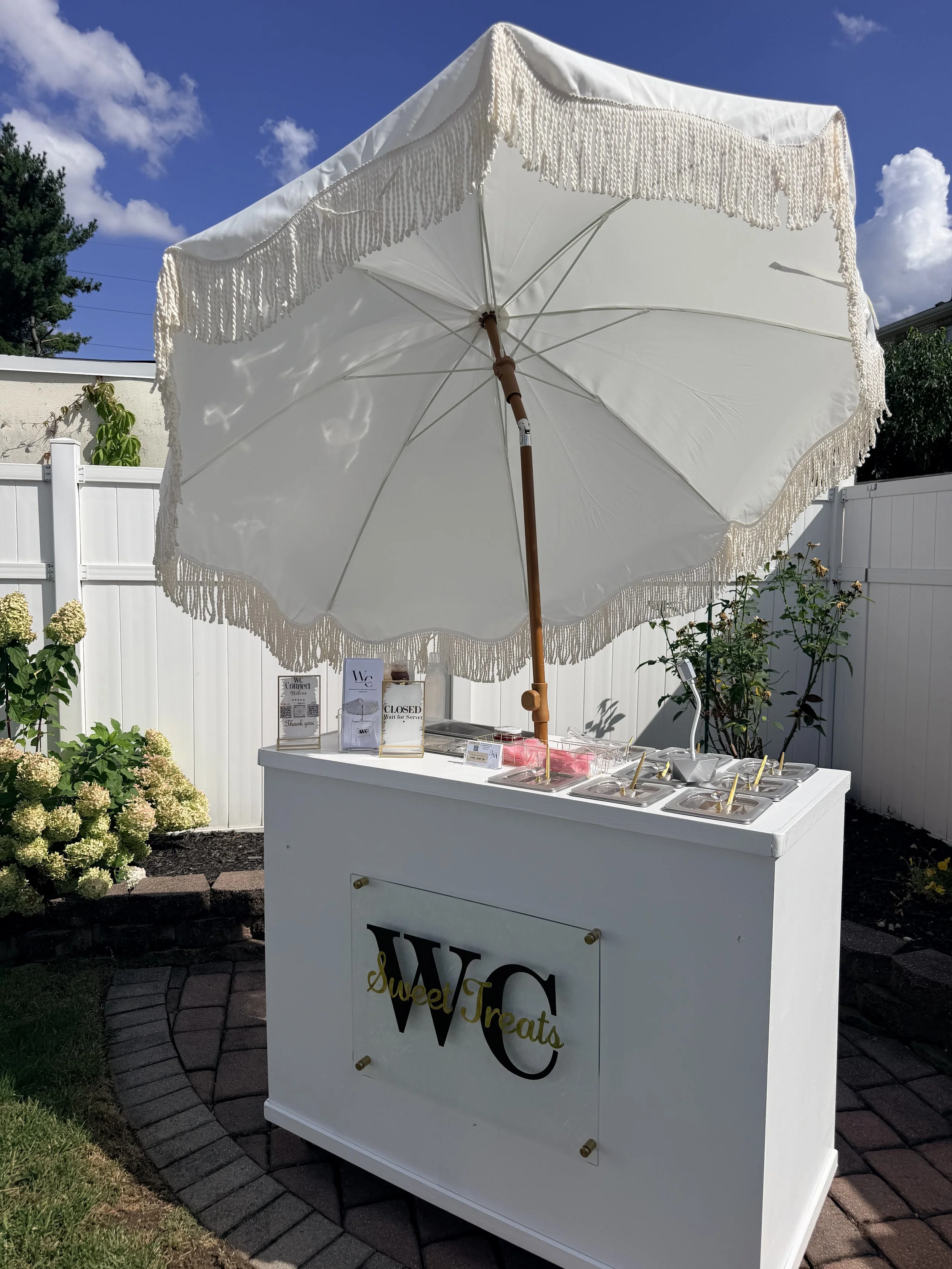 A white ice cream stand with an umbrella, decorated with fringe, outdoors under a blue sky with clouds. The stand has signs and containers for ice cream and toppings, and a white fence in the background.