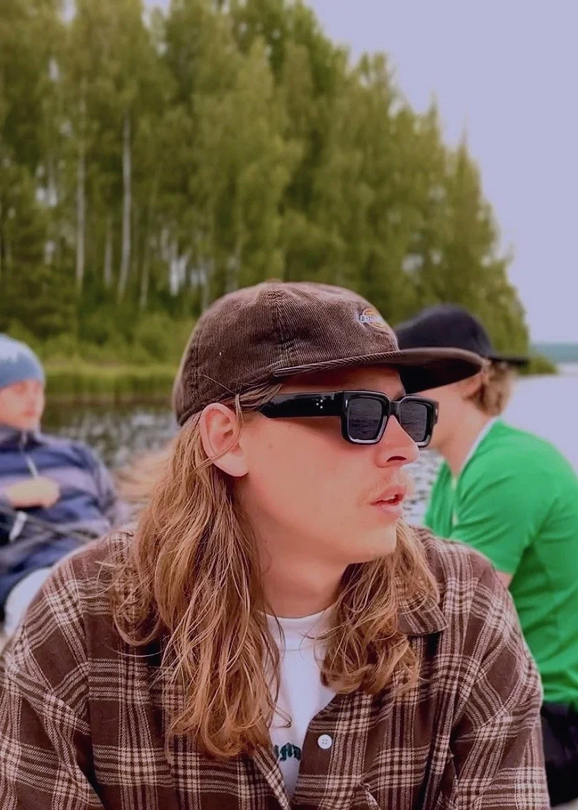A man with long wavy hair wearing a brown baseball cap, black sunglasses, and a brown plaid shirt outdoors near a body of water with trees in the background.
