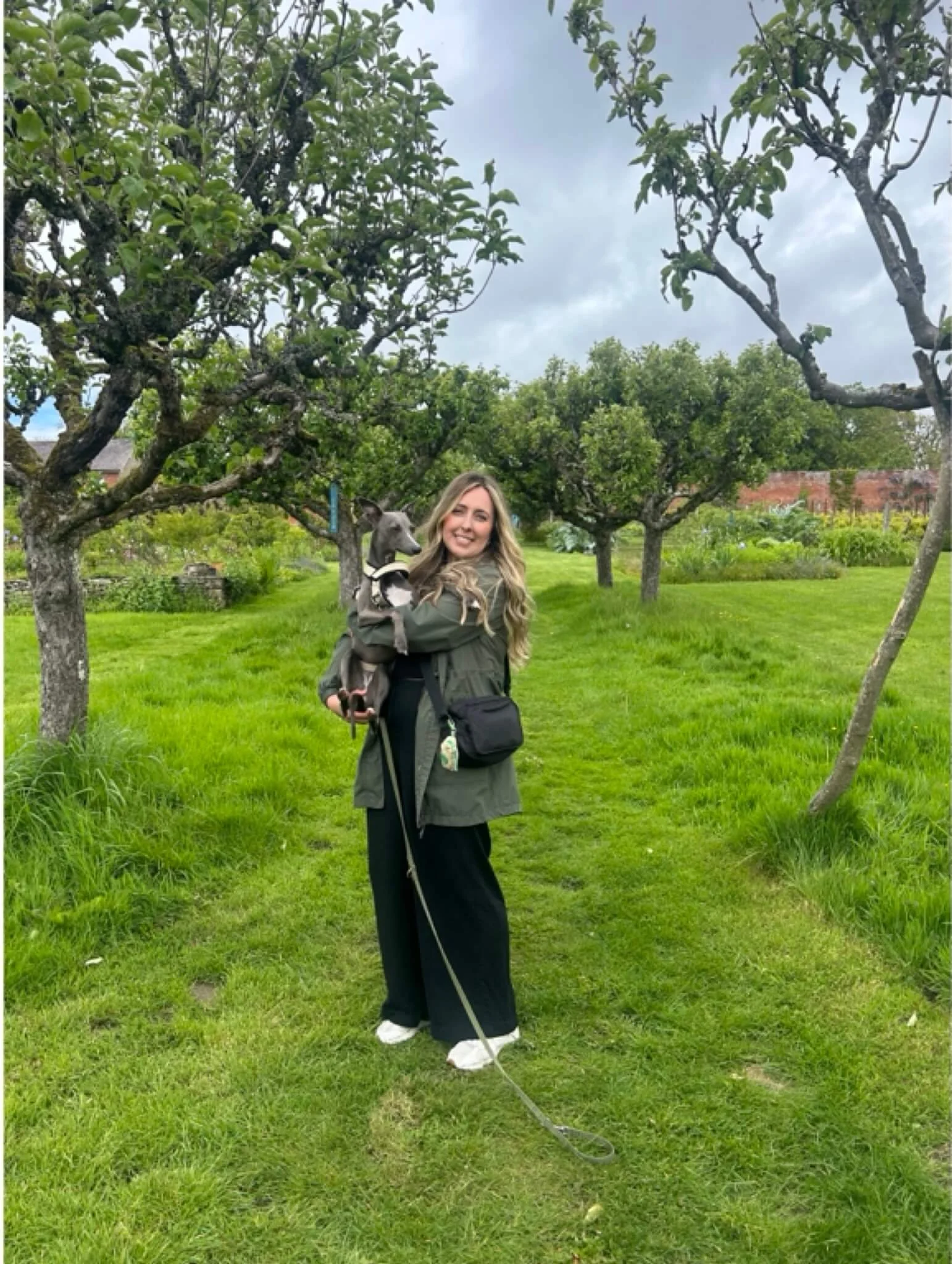 Amy James artist, picture book illustrator, smiling and holding a small dog in a lush green garden with trees and a cloudy sky.