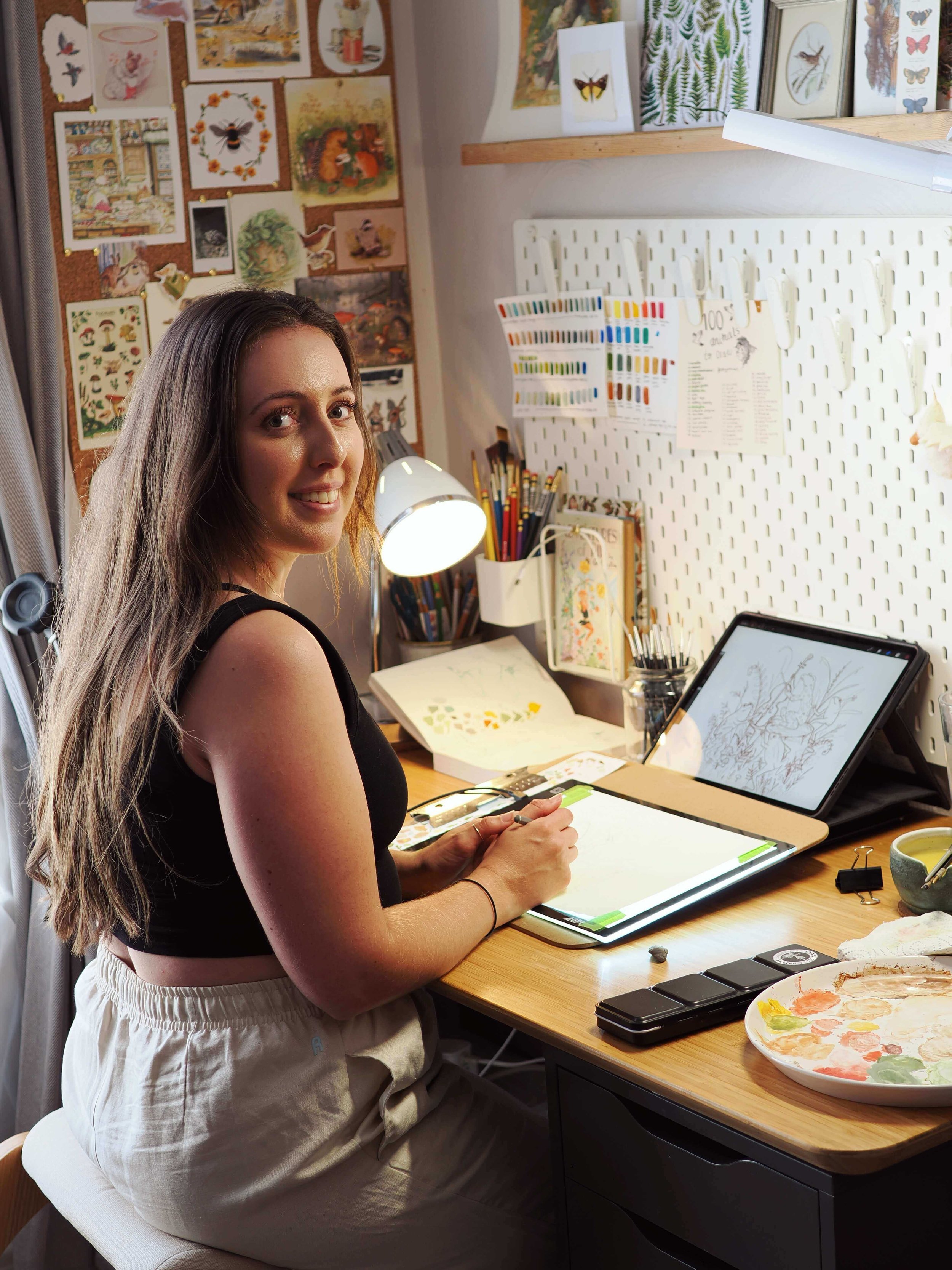 Amy James sitting at a desk with art supplies, watercolour paints, and a sketch. Behind her, a wall is decorated with various art and nature illustrations.