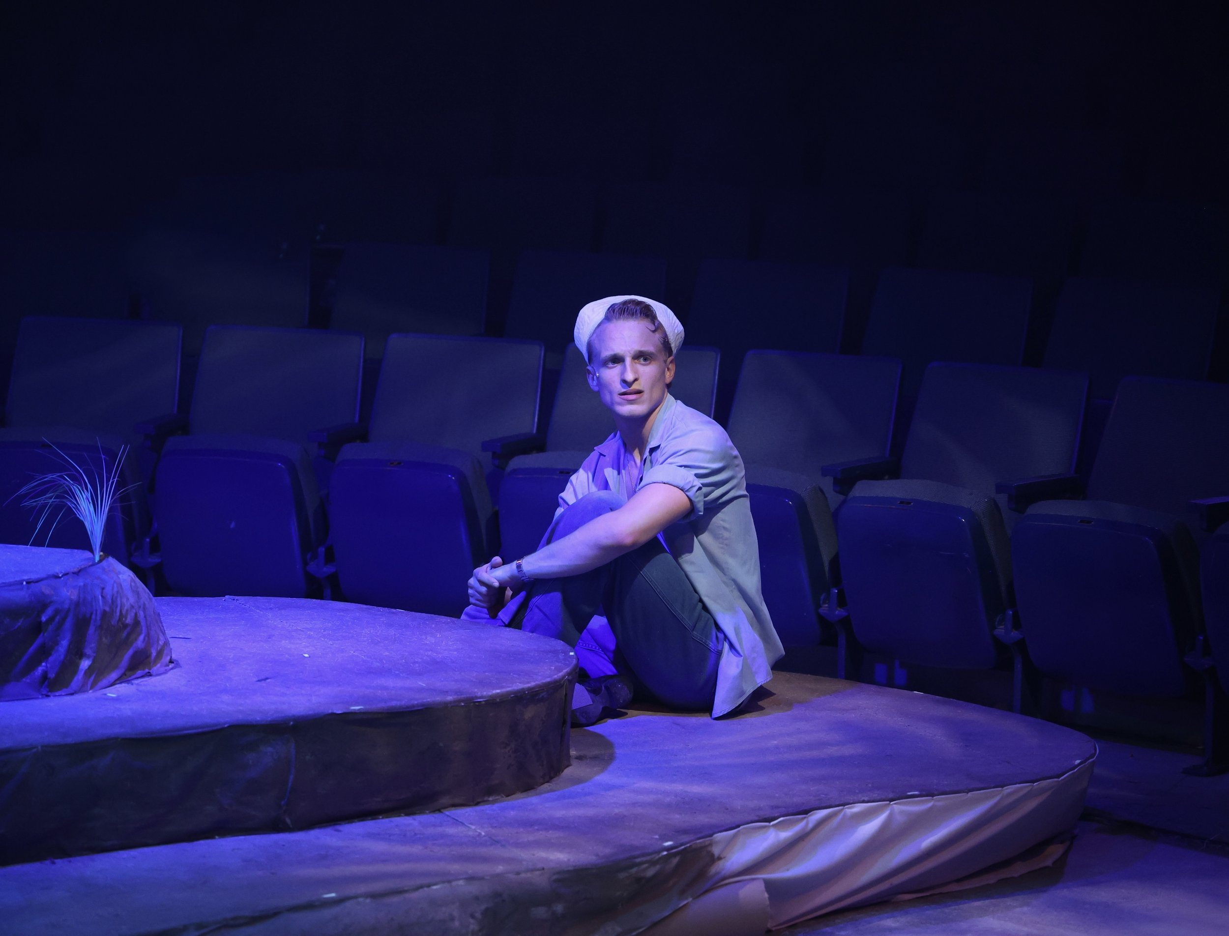 A young man with light skin, sitting on the floor of a theater stage, looks serious. He is wearing a gray jacket, black pants, and a white hat. The stage has a round platform with a faux plant, and empty theater seats are visible in the background under blue lighting.