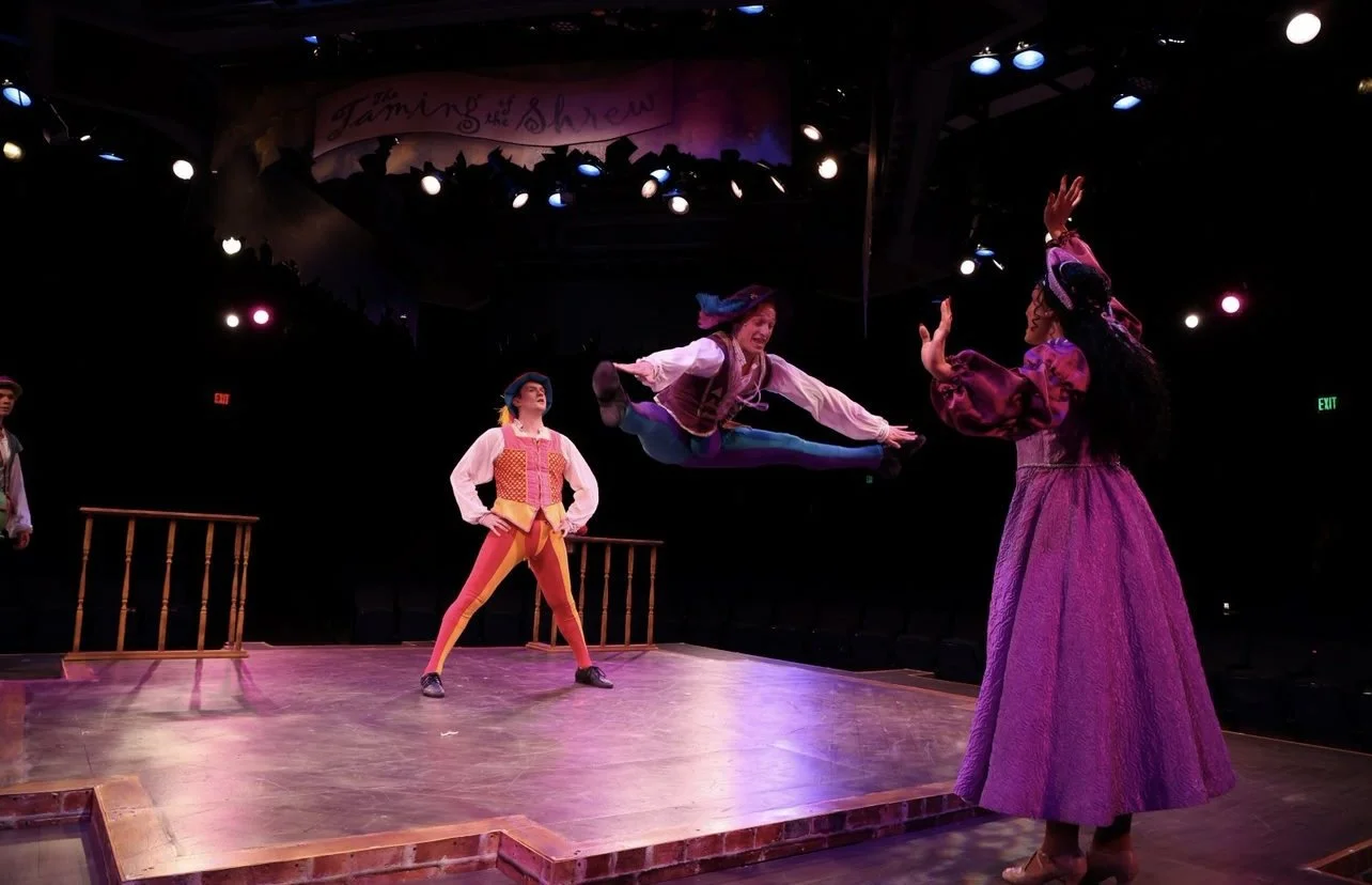 The image shows a theatrical performance titled 'Jaming of the Show.' Three actors in colorful costumes are on a stage with a wooden floor. One actor is mid-air doing a jump, while the other two are standing and watching. The background is dark with stage lighting.