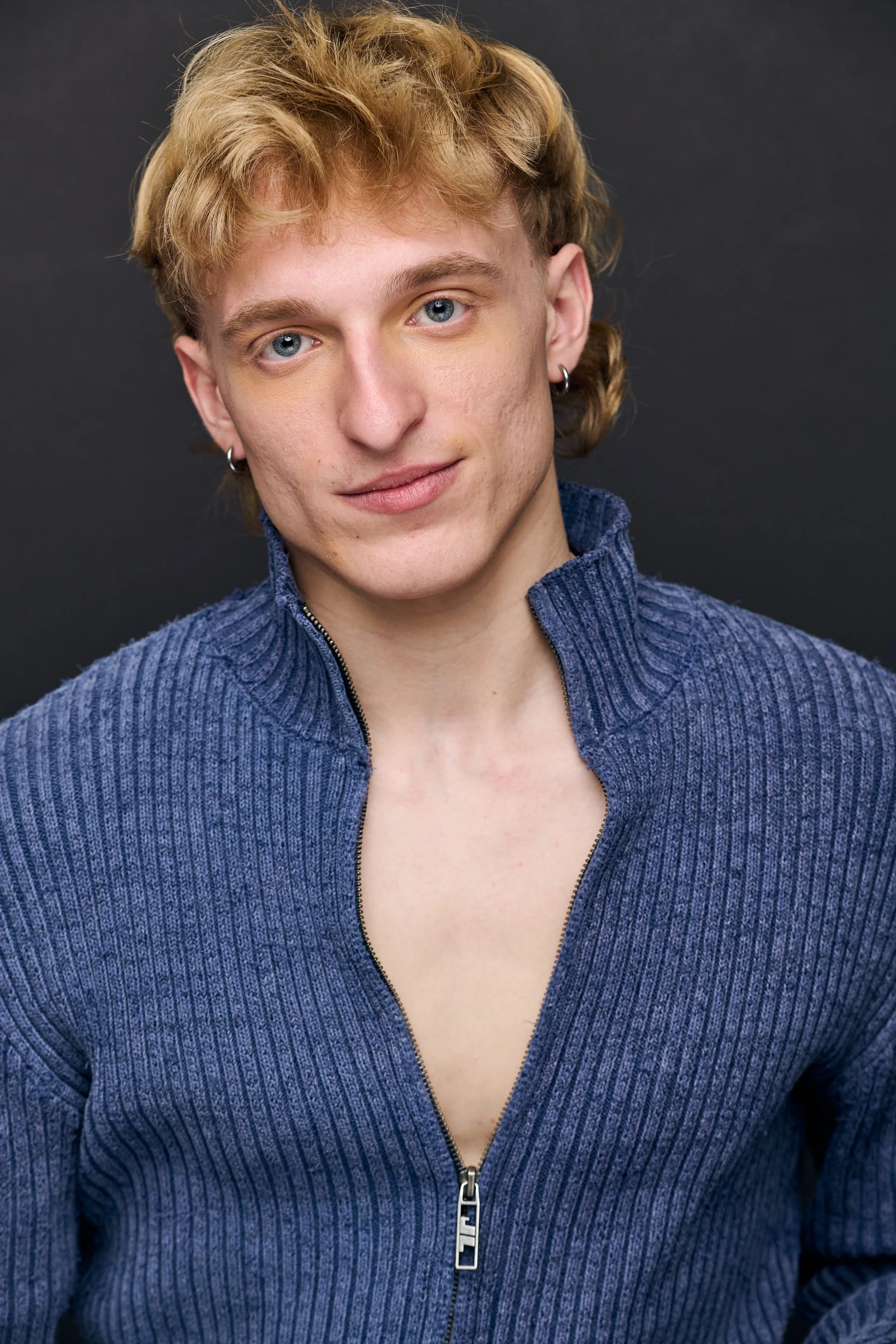 A young person with blond curly hair, blue eyes, and earrings, wearing a blue zip-up sweater, posing against a dark background.