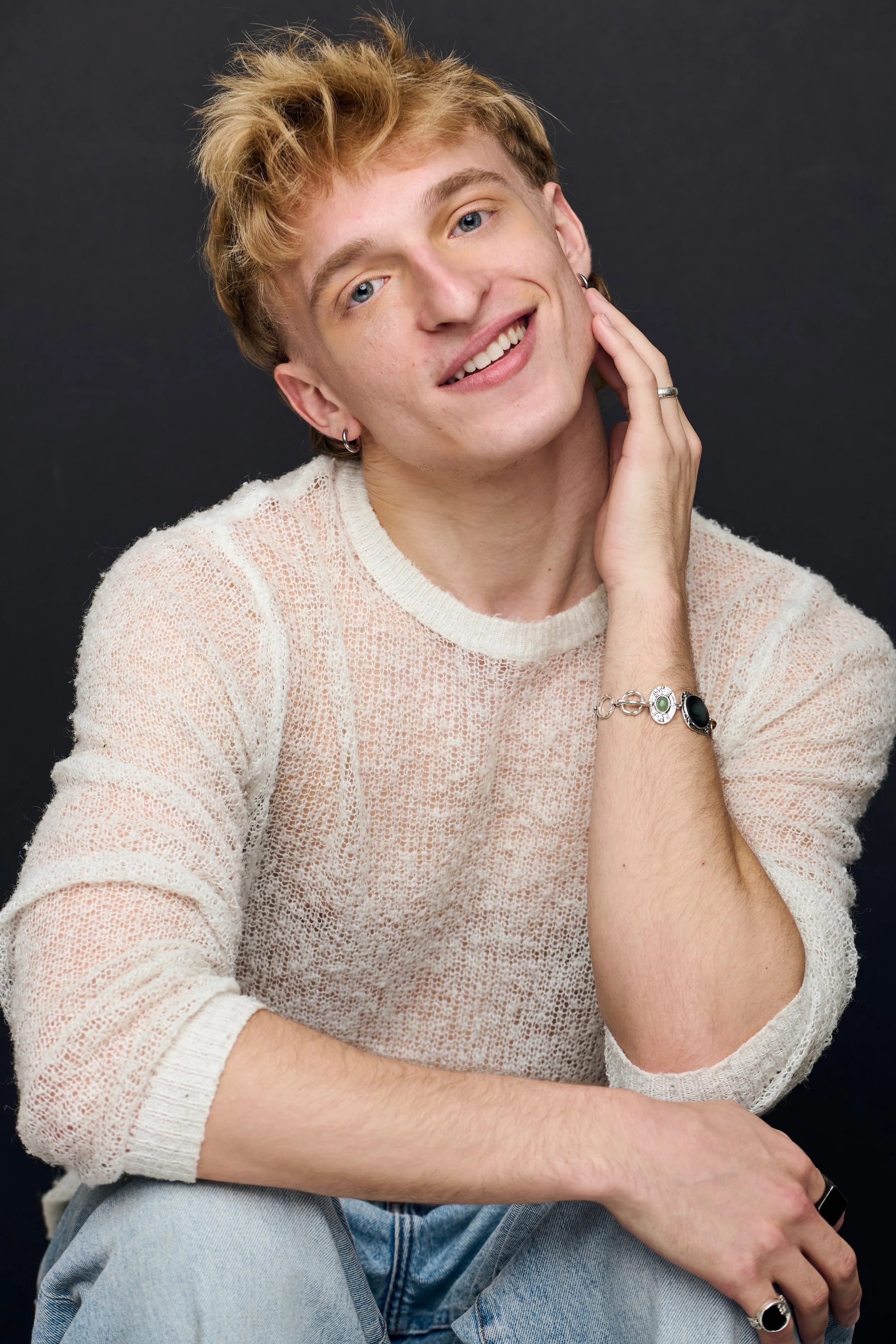 A young man with blond hair and blue eyes posing against a black background. He is smiling and slightly tilting his head, touching his face with his right hand. He is wearing a see-through, off-white knitted sweater, light blue jeans, earrings, and bracelets.