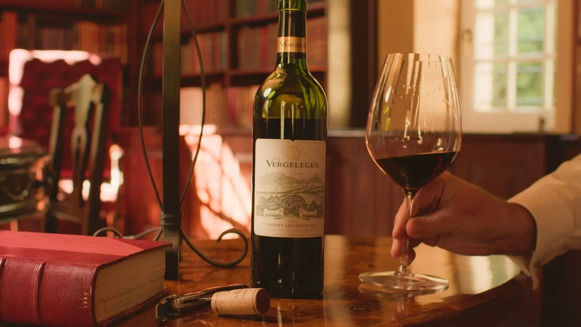 Vergelegen Winemaker — next to a bottle of Vergelegen Cabernet Sauvignon 2021 on a wooden table inside a cozy, book-filled room.