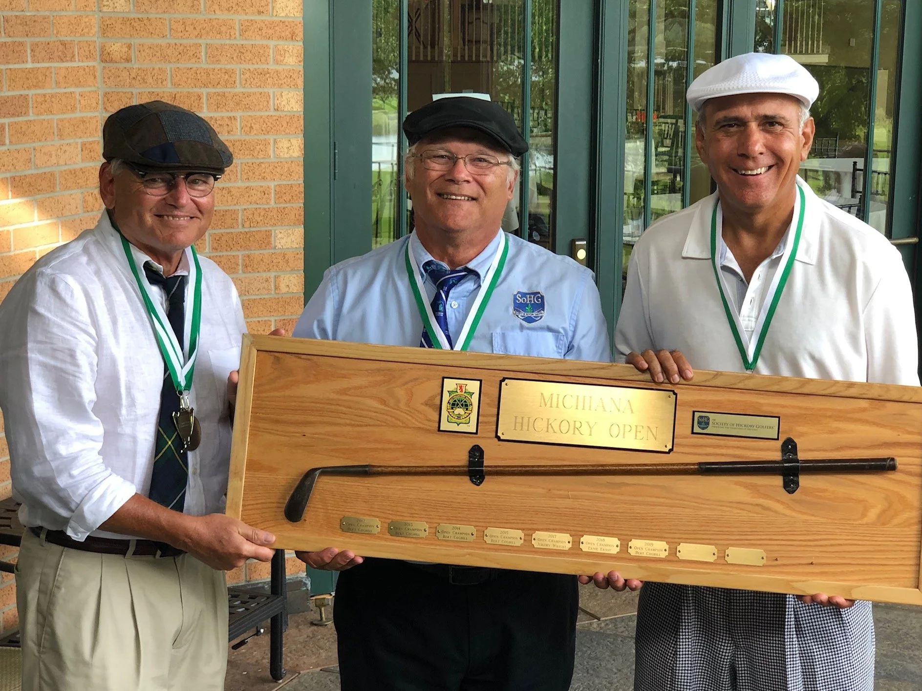 Three men wearing hats and medals around their necks, holding a large wooden plaque with inscriptions and small plaques attached at the bottom, standing outdoors in front of a brick wall and glass doors, smiling at the camera.
