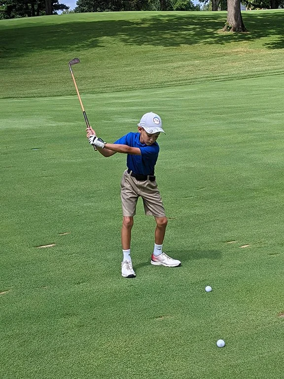 A young boy wearing a white cap, blue polo shirt, beige shorts, and white shoes scored with red accents, is preparing to hit a golf ball on a green golf course. He is holding a golf club and standing next to two golf balls on the grass, with trees and a slightly hilly terrain in the background.