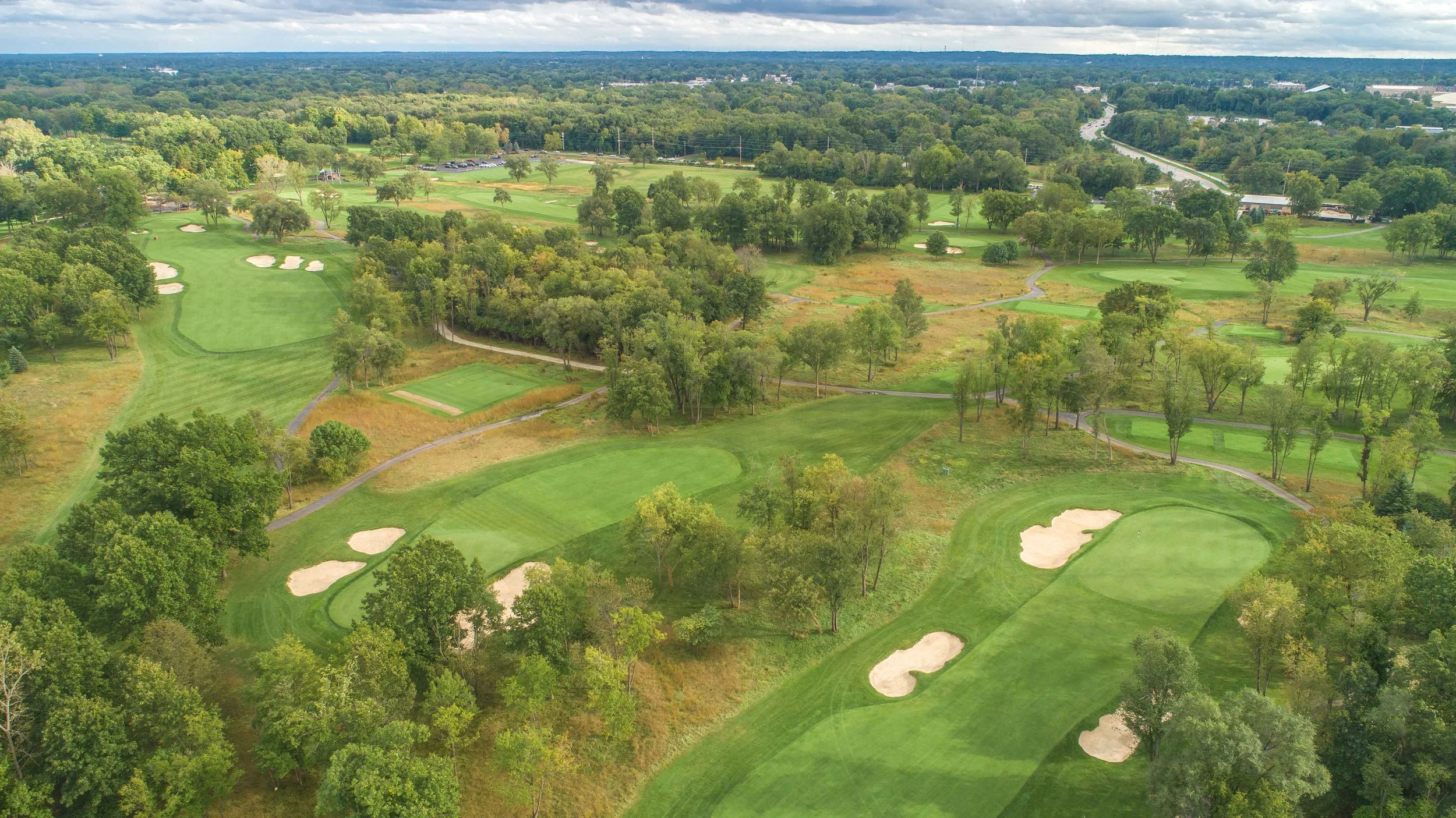 An aerial view of a golf course surrounded by trees, with multiple sand bunkers, green fairways, and pathways, extending towards a city area in the background under a partly cloudy sky.