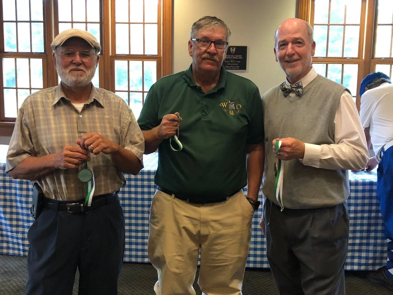 Three men standing indoors near a window, holding medals, in a setting that appears to be a golf course event or ceremony.