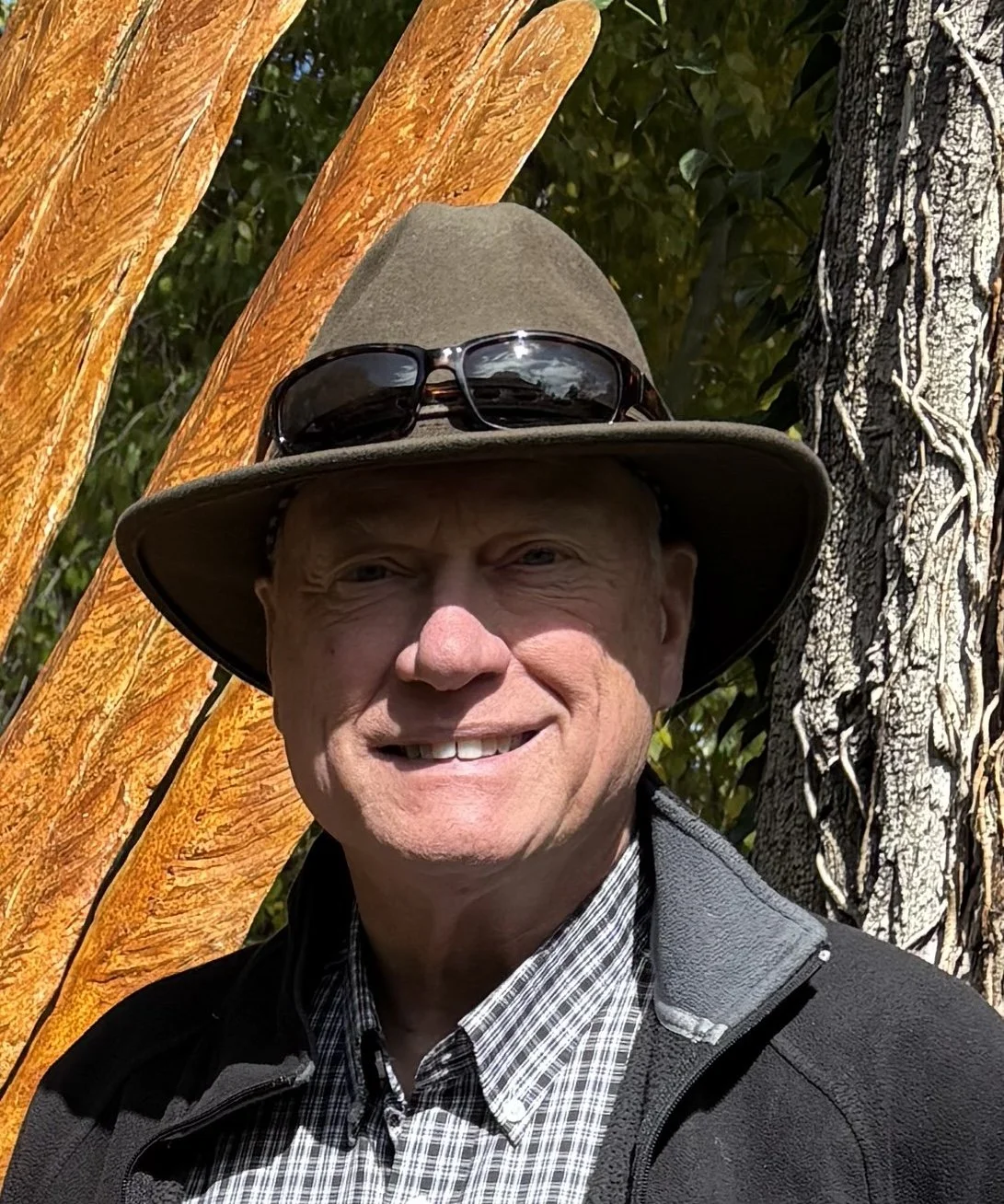 A man wearing a light brown hat, gloves, and a dark shirt is outdoors in a grassy field, holding a large white bag filled with plant debris or weeds, smiling.