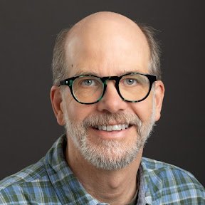 Smiling man with glasses, a gray beard, wearing a dark green baseball cap and a blue T-shirt, standing outdoors near houses and plants, under cloudy sky.