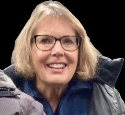 Young woman with shoulder-length black hair, smiling, wearing a blue top, outdoors with green foliage in the background.