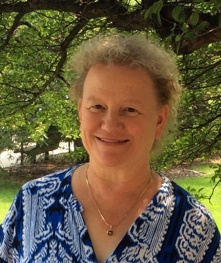 A group of three people standing on a rocky shoreline near a boat on a calm lake, with one person pointing towards the water, and trees in the background.