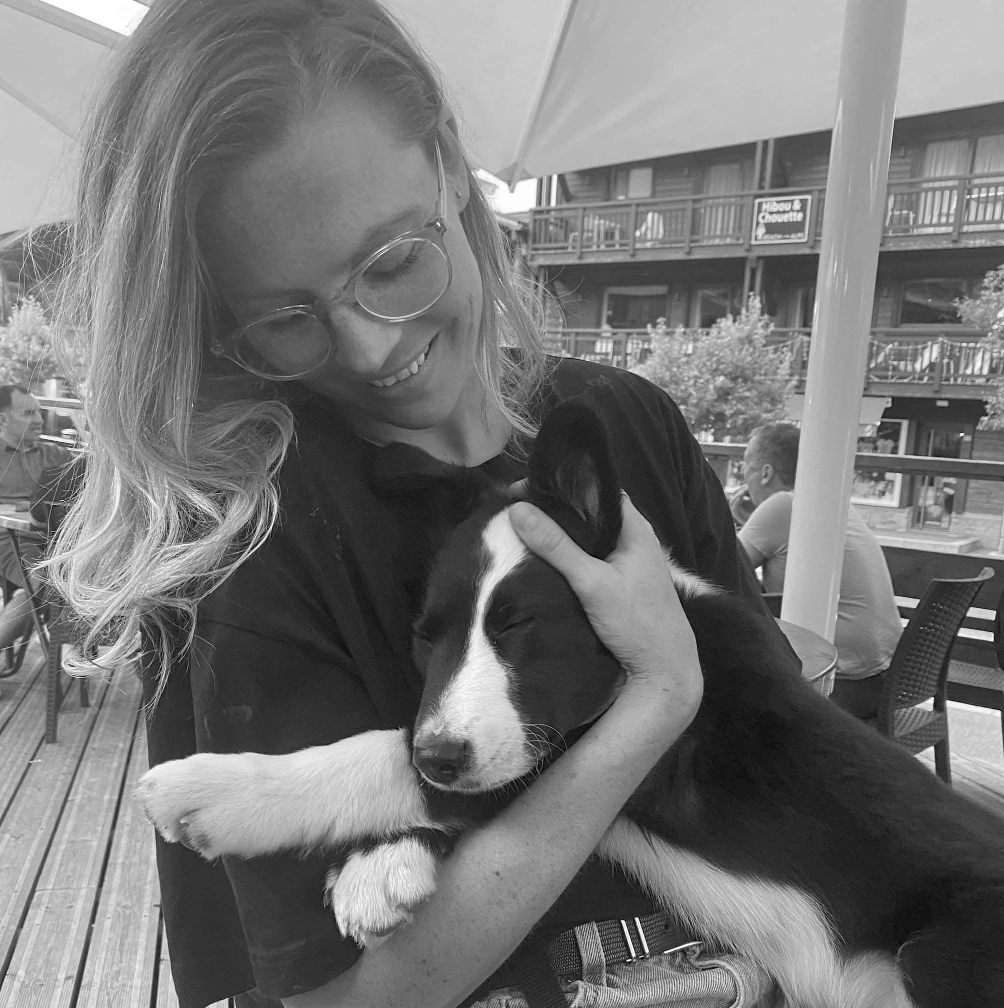A woman with glasses smiling and holding a sleeping puppy in her arms, outdoors on a patio.