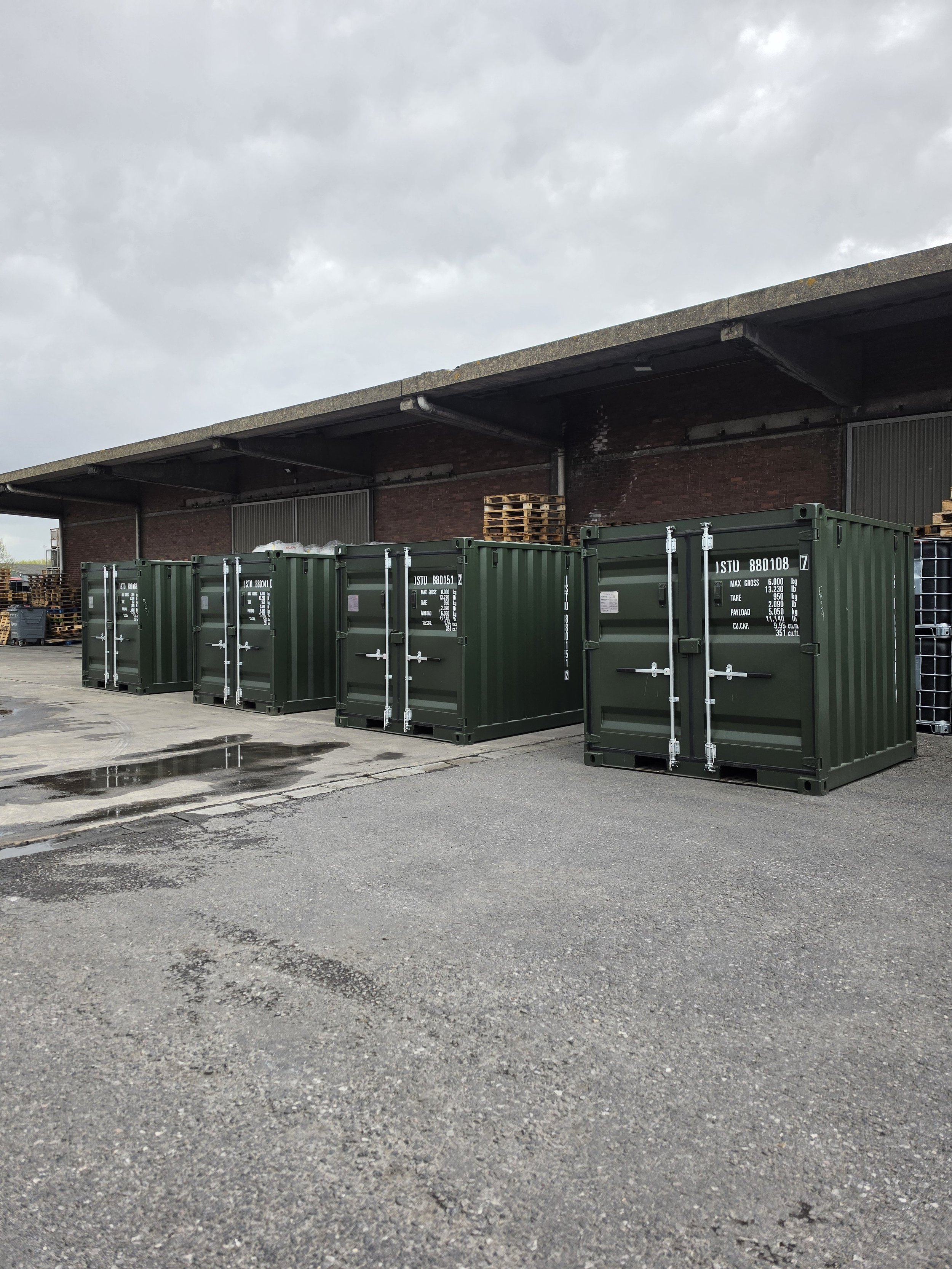 Green storage containers lined up outdoors under a cloudy sky, with pallets and scattered water puddles on the concrete ground.