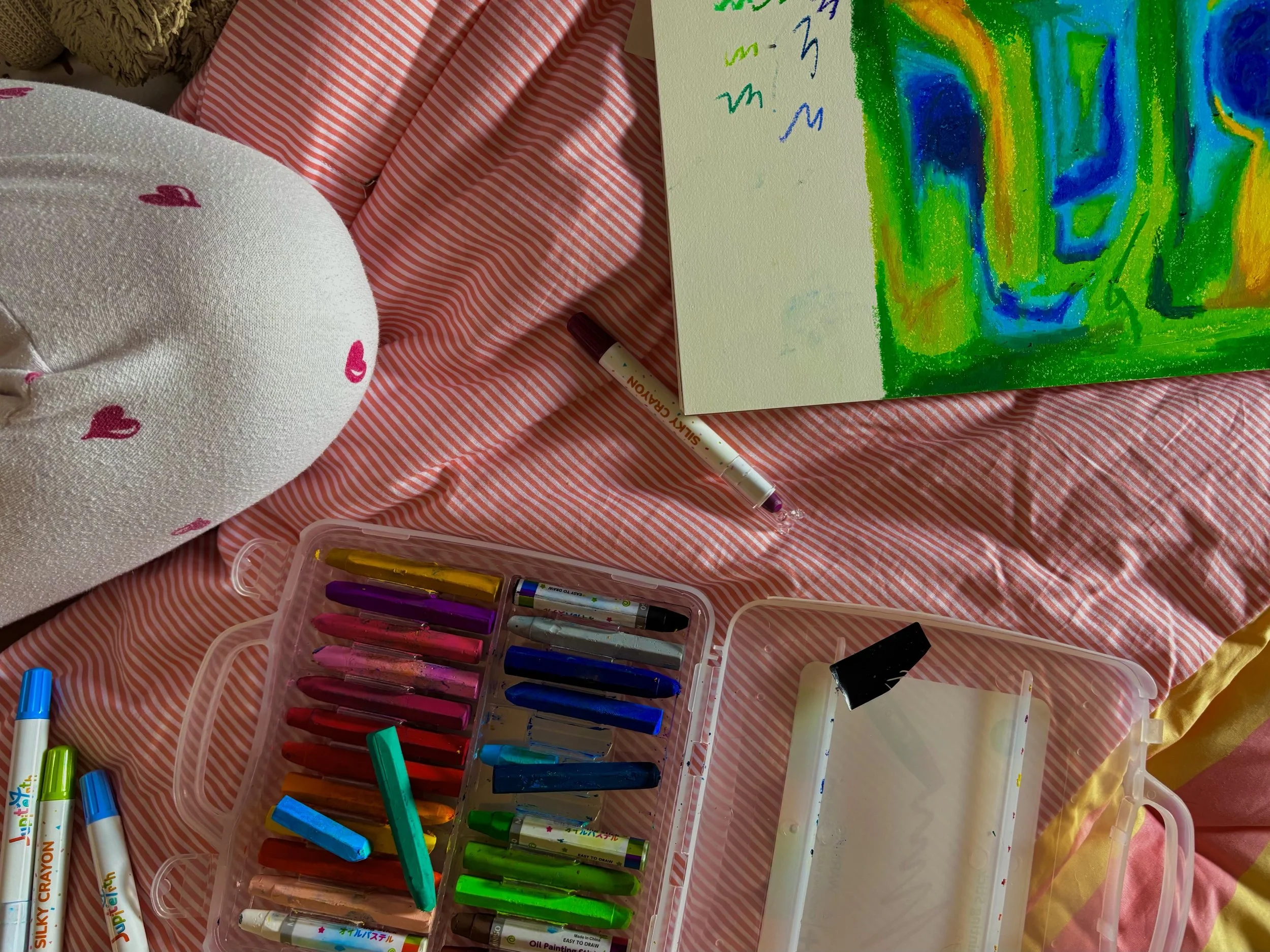 A pink striped bedsheet with art supplies including a white plush heart with pink hearts, a case of colorful gel pens, and a sketchbook with a partially finished colorful abstract drawing and some scribbles.