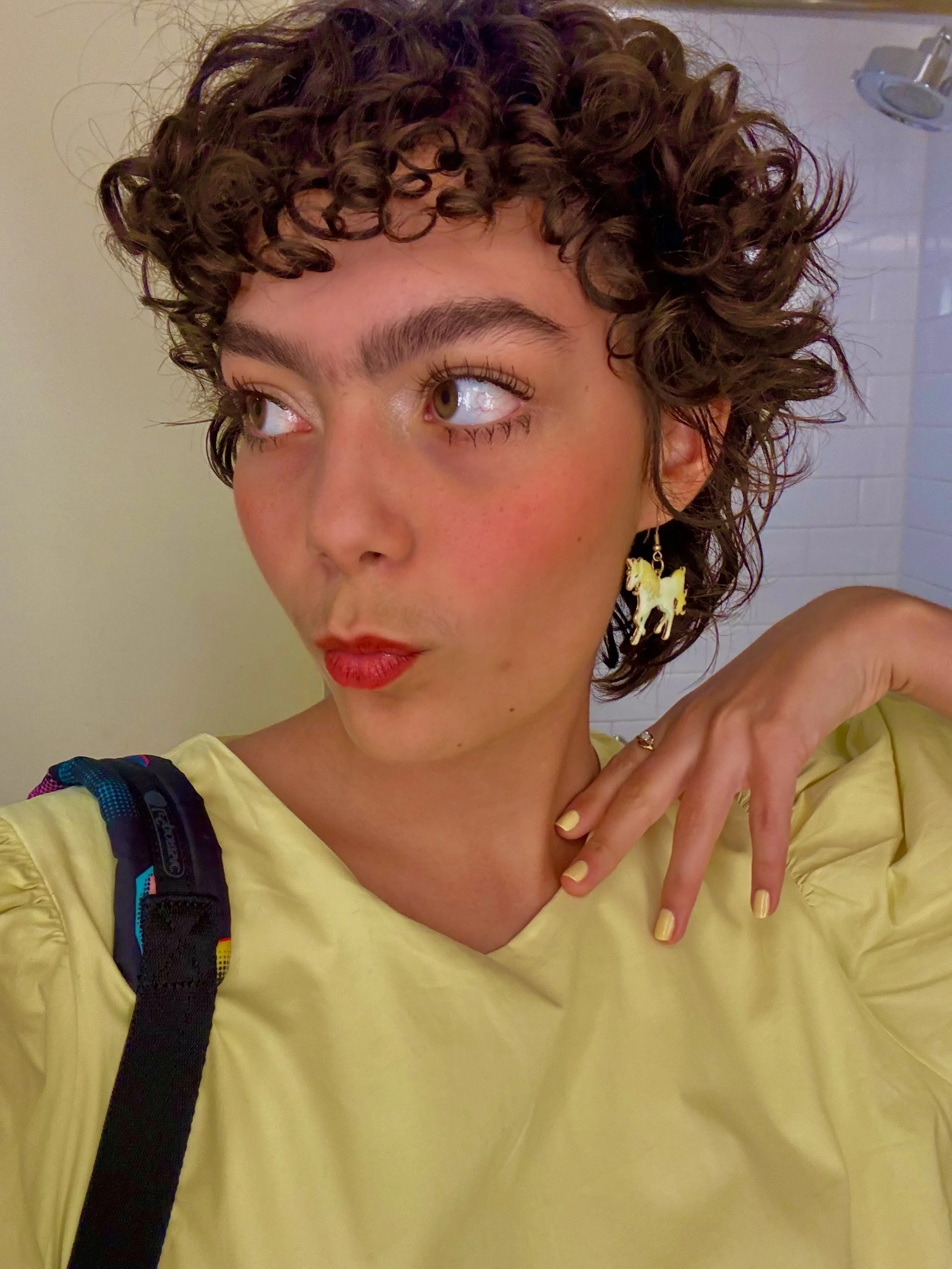 Esther wearing bright red lipstick, yellow eyeshadow, and a yellow top, with a unicorn earring on one ear. She has a confident expression, with one hand raised near her neck and her lips making a kissing gesture.