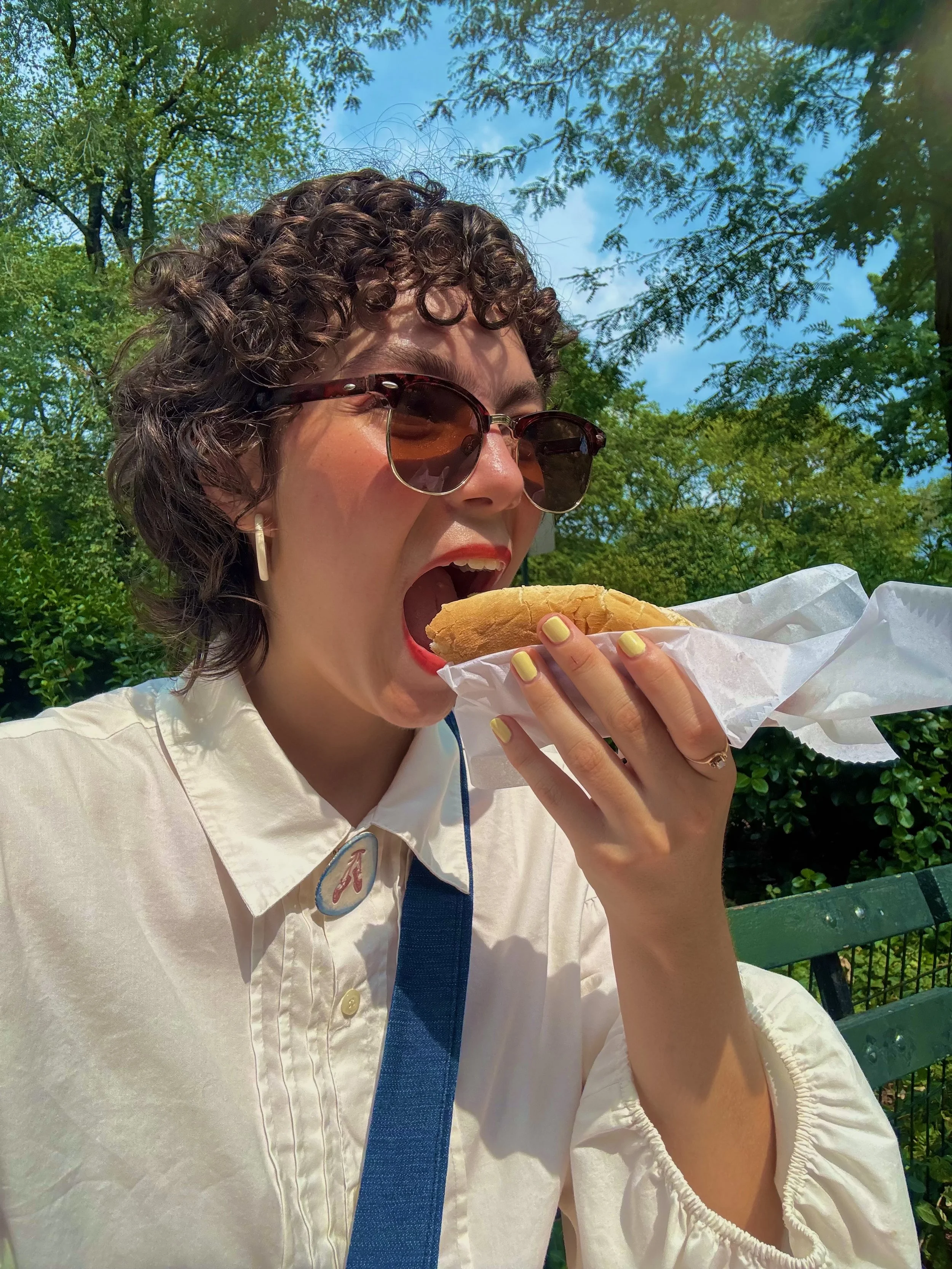 Esther wears sunglasses and a white shirt with a pointe shoe pin while eating a hot dog outdoors on a sunny day at central park in New York City.