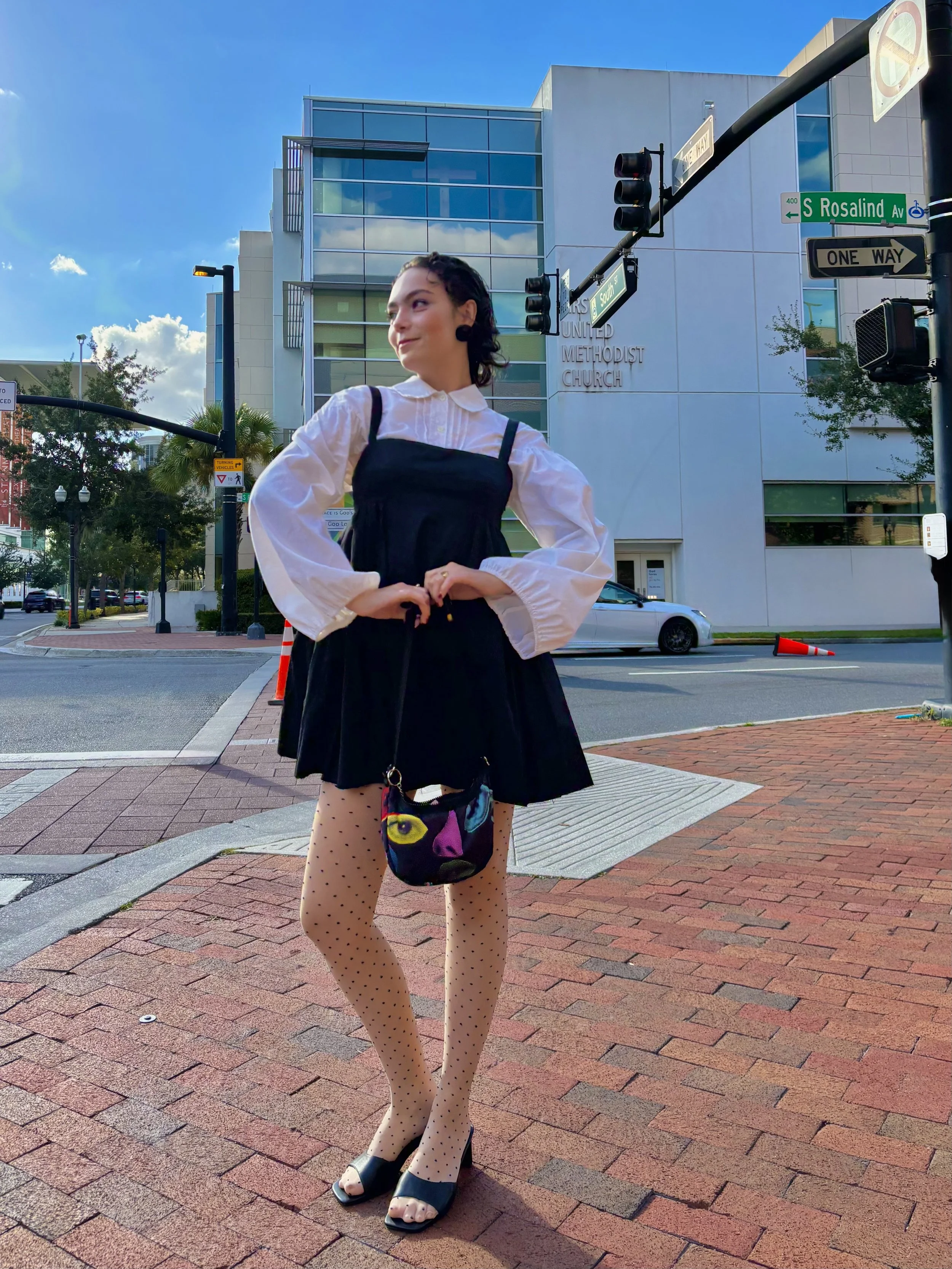 Esther stands at an intersection in front of a city Church posing for a picture while wearing a white blouse under a black dress, polka dot tights, black heels, carrying a colorful bag.