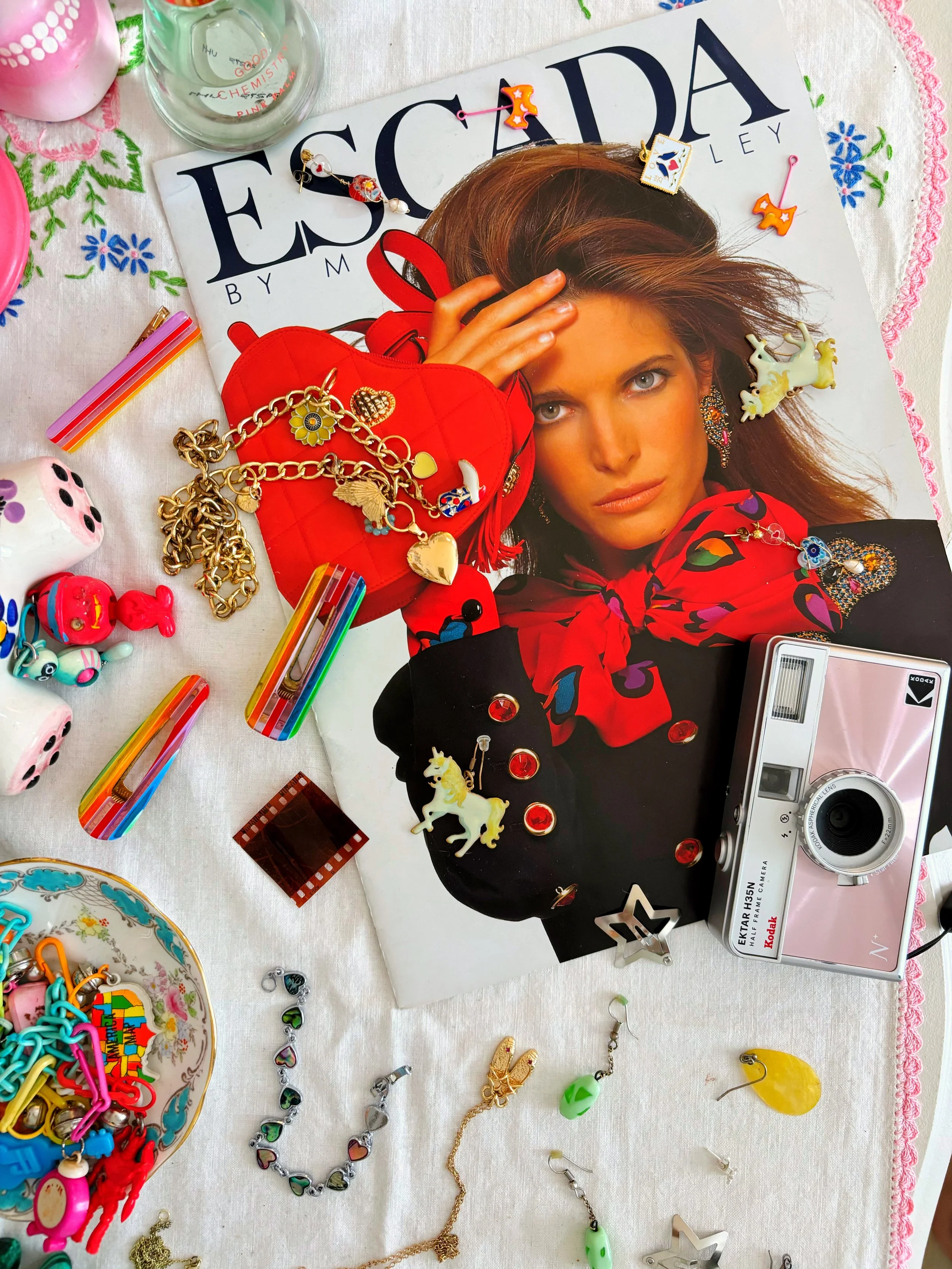 A colorful flat lay of jewelry, toys, and accessories around a magazine featuring a woman with brown hair. Items include a red heart-shaped purse, gold chains, charms, toys like a unicorn and pig figures, rainbow-colored hair clips, earrings, a camera, and a bowl of colorful clips and toys on a white and pink embroidered tablecloth.