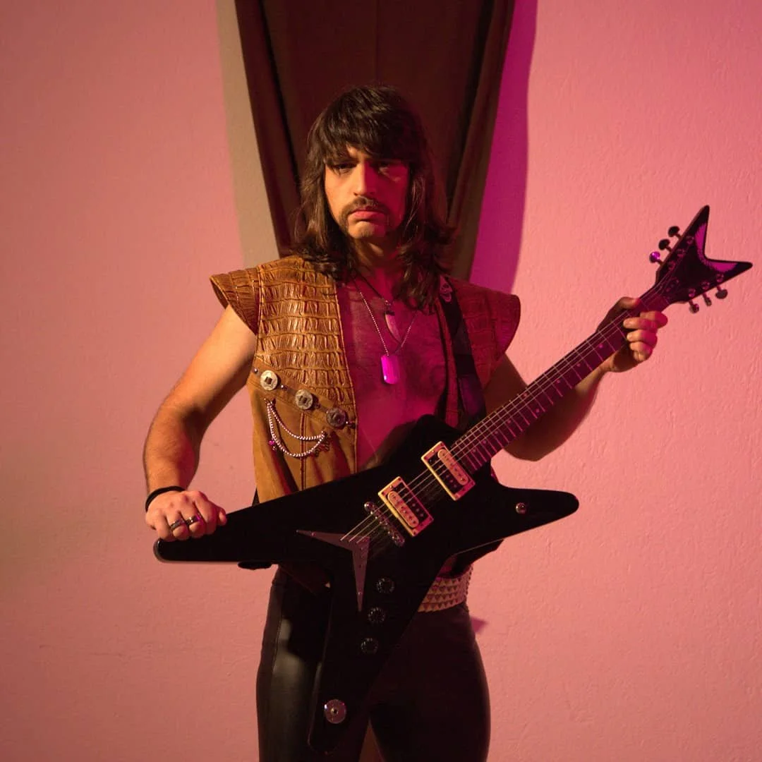 Person with long dark hair wearing a sleeveless brown vest holding a black electric guitar.