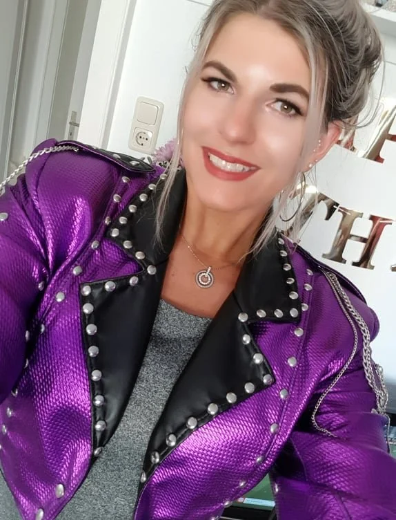 A woman with blonde hair tied back, wearing a purple metallic jacket with silver studs and a black lapel, a gray top, hoop earrings, and a silver necklace, smiling indoors.