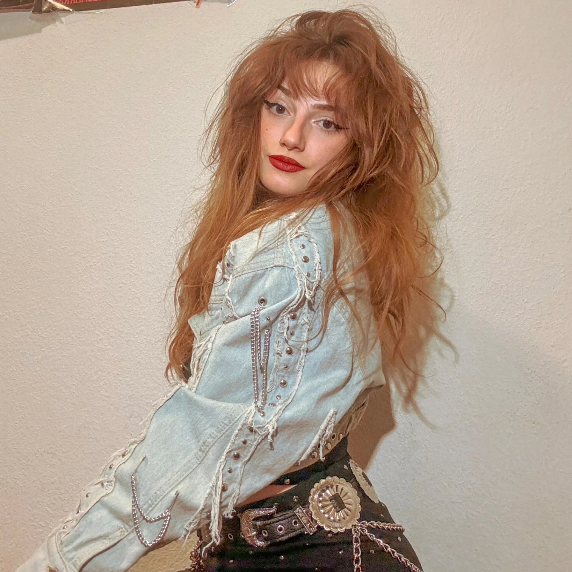 Young woman with long, red, curly hair wearing a distressed denim jacket with silver embellishments and chains, and black pants with a decorative belt, standing against a plain wall.