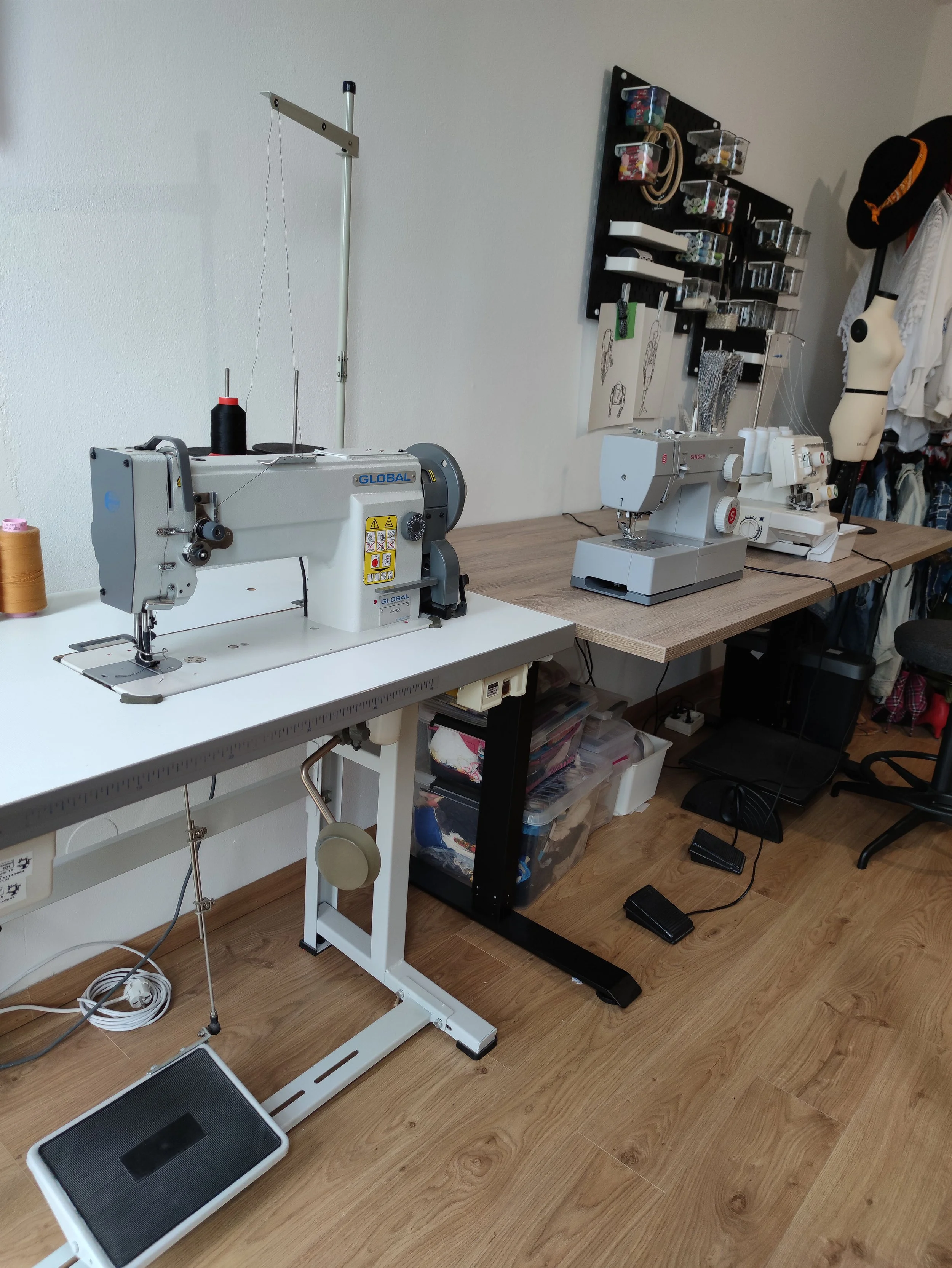 A sewing workspace with two sewing machines, one white and one gray, placed on separate tables. There is a foot pedal on the floor connected to the machines, and various materials such as thread, fabric, and tools are visible. A wall-mounted organizer holds sewing supplies, and there are sketches pinned to the wall. A mannequin dressed in white and a rack with clothing items are partially visible on the right side.