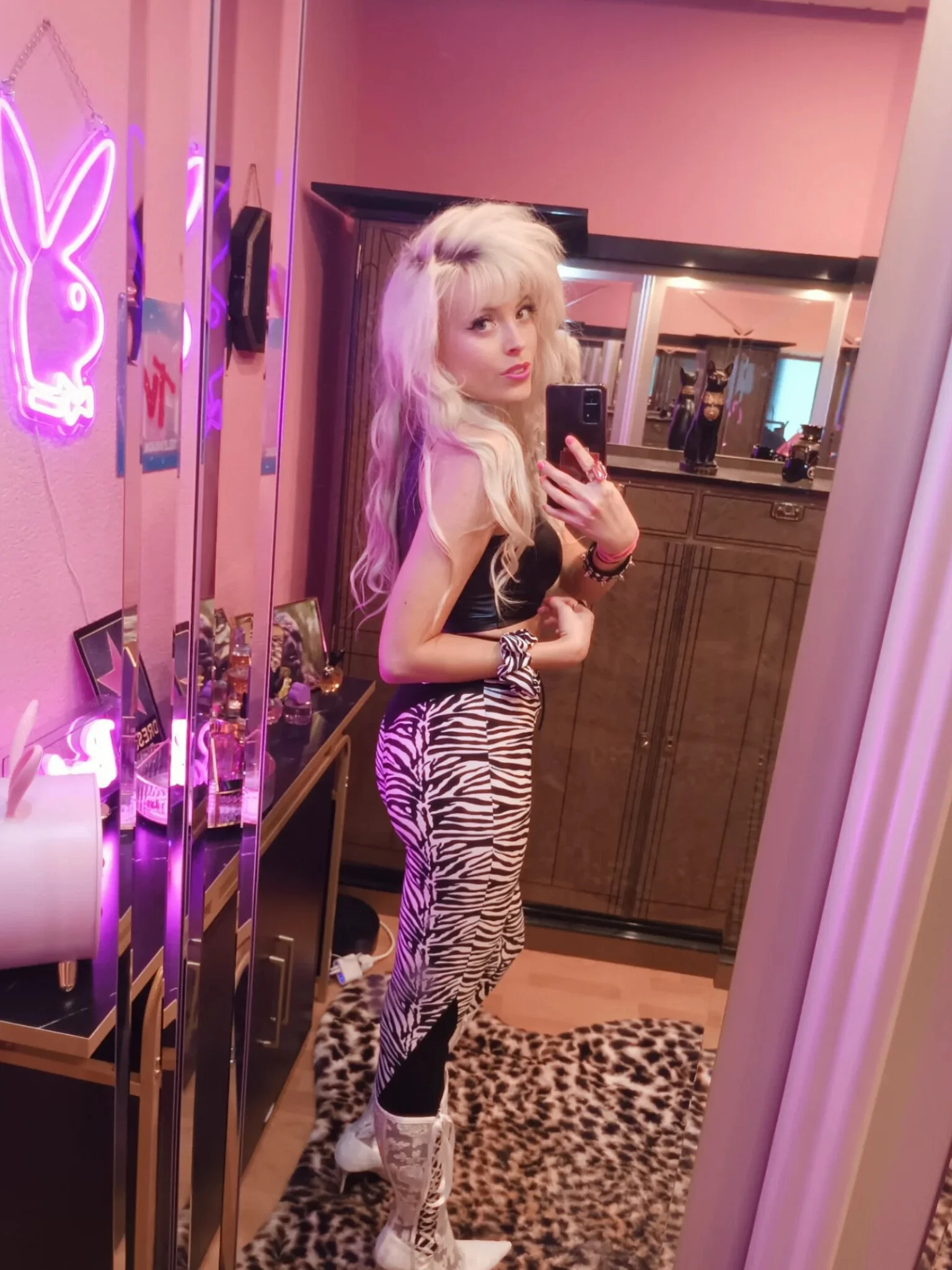 A woman taking a mirror selfie in a room with pink lighting and neon bunny shaped signs. She has long blonde hair, wears animal print zebra striped leggings, white high heels, and a black top.