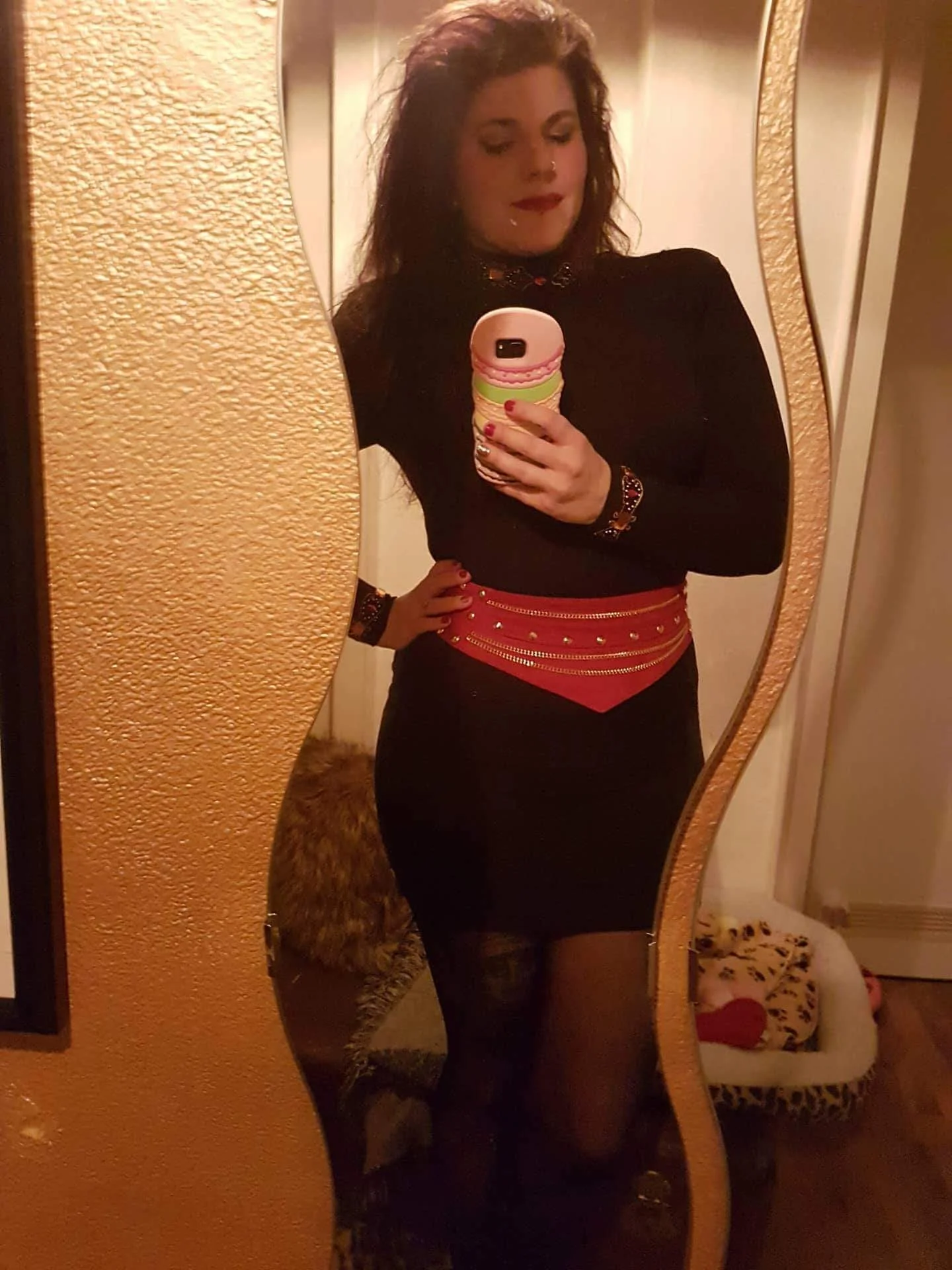 A woman takes a mirror selfie, wearing a black long-sleeve top, a red belt with gold chains, black tights, and holding a phone with a pink, green, and cream-colored macaroon-ice cream shaped phone case. She has long hair, red lipstick, and is in a ro