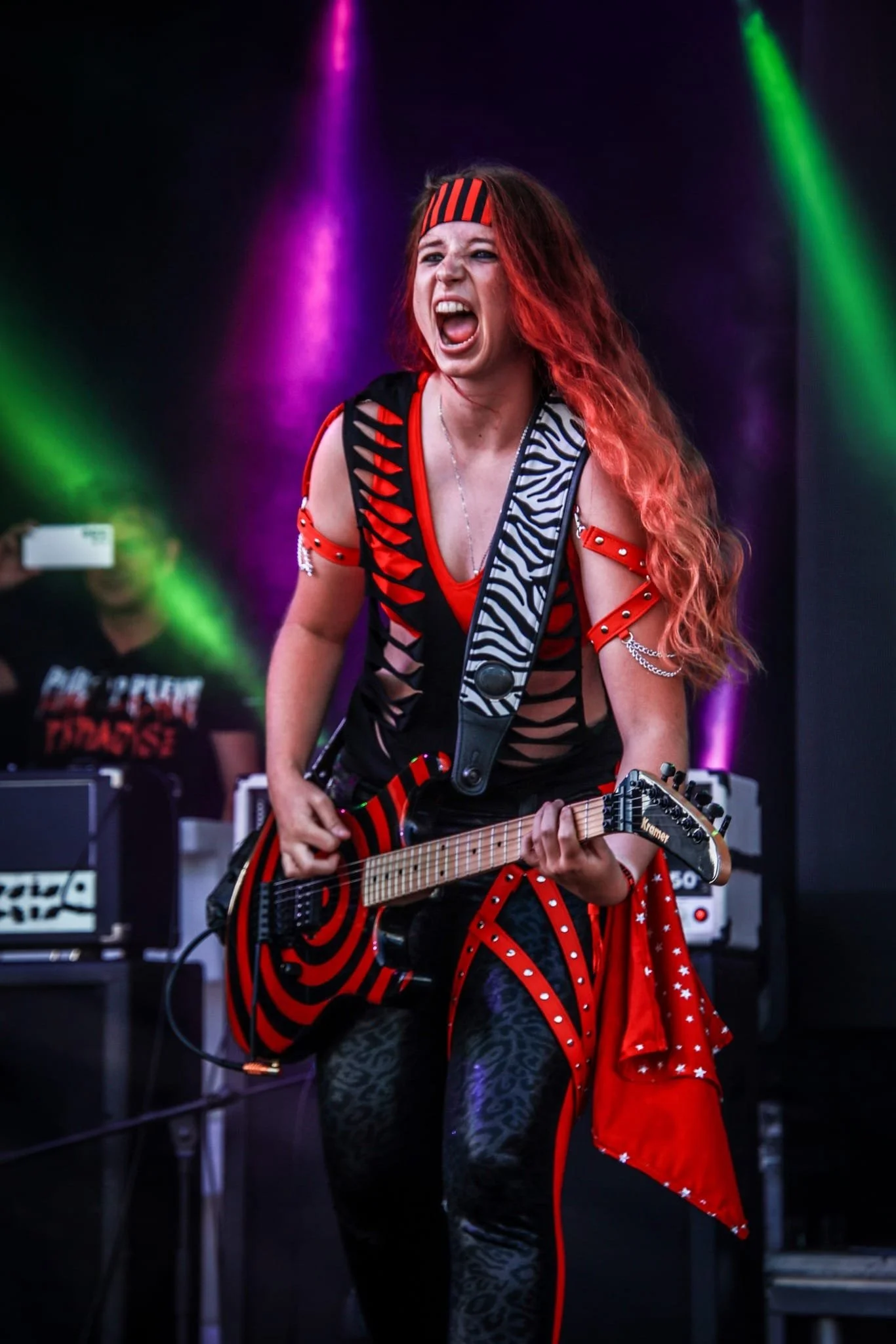 A female guitarist with long red hair passionately performing on stage, wearing a black and red striped headband and a black top with red and white accents, surrounded by colorful stage lights.