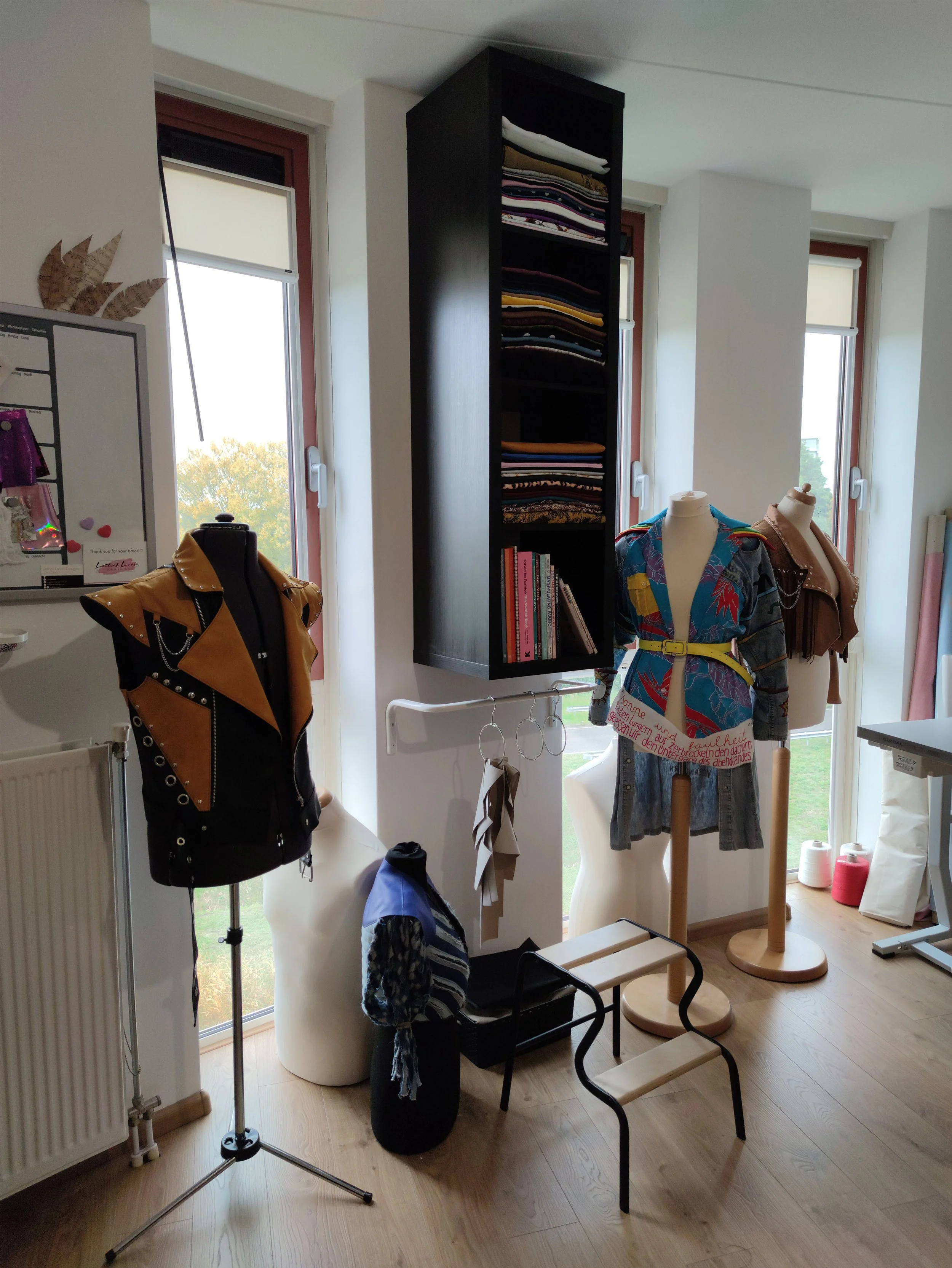 A room with three large windows, garden views, and mannequins with clothing on display. There is a black shelving unit with folded clothes, a wooden chair, a backpack, and a clothing rack. The floor is wooden, and there are various objects like a mannequin, books, and decorative items.