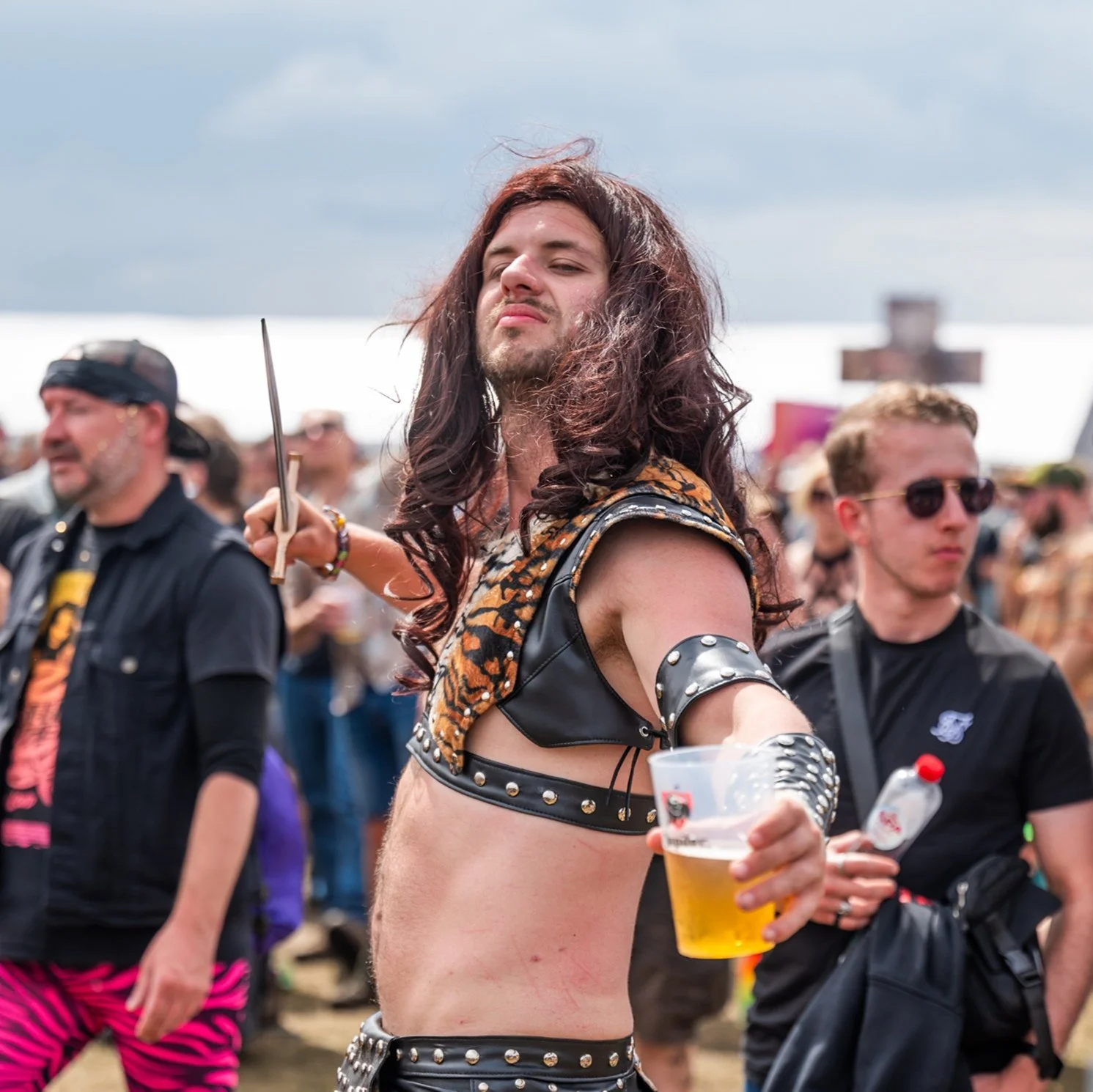 Person with long hair and punk-style leather outfit holding beer at outdoor music festival.