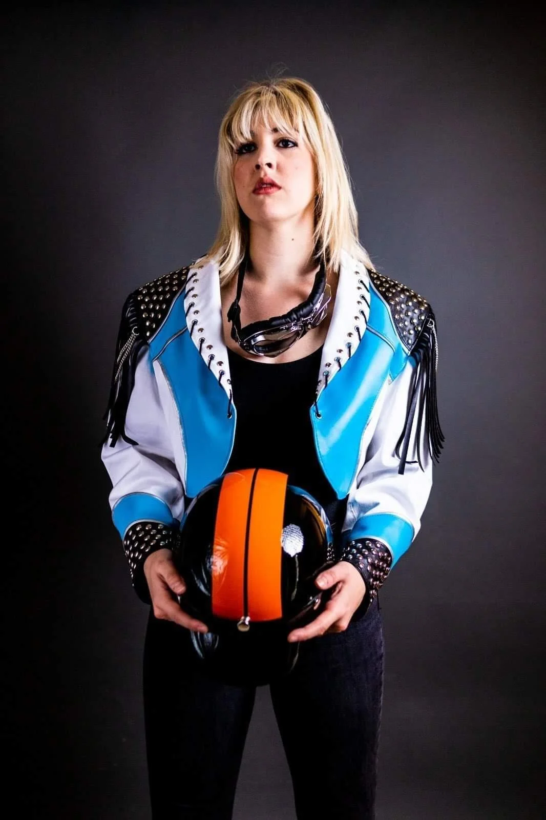 A woman with blonde hair wearing a black shirt and a blue and white jacket with studded black leather accents, holding a motorcycle helmet with orange and black stripes, standing against a dark background.