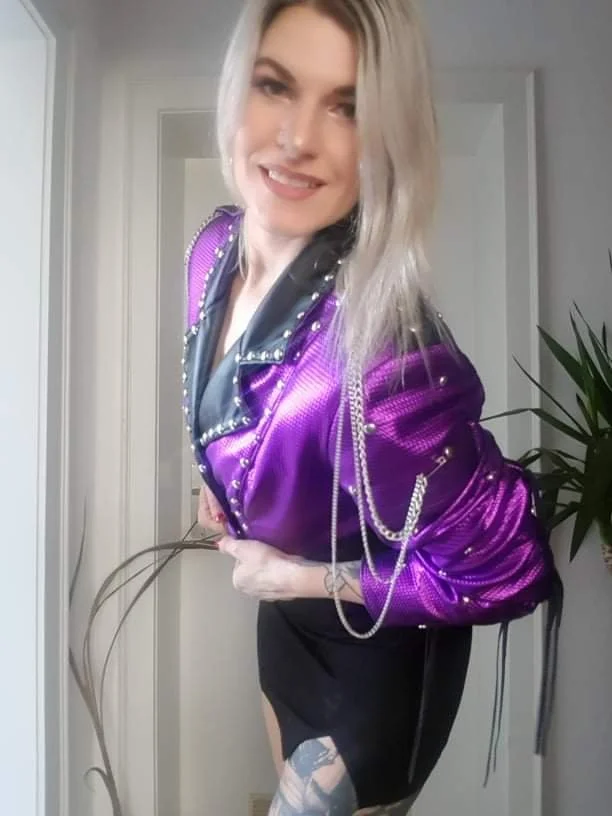 A young woman with blonde hair smiling at the camera, wearing a shiny purple jacket with silver chains and studs, and a black skirt, standing indoors near a plant.