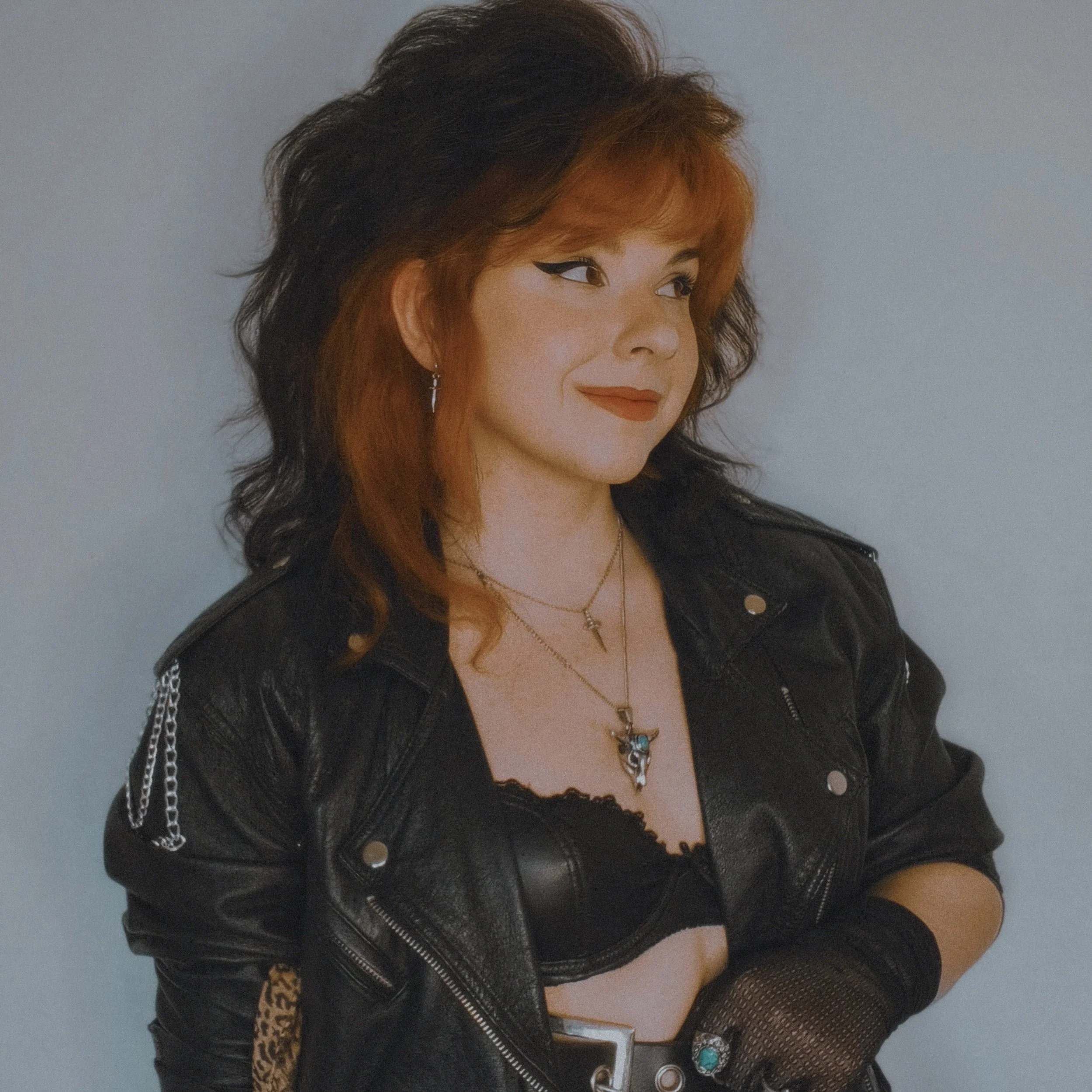 Zora Clevers, the owner of Lethal Lover Designs. A woman with red and black hair wearing a black leather jacket, black fishnet gloves, layered necklaces, and makeup with winged eyeliner and red lipstick, posing against a light gray background.