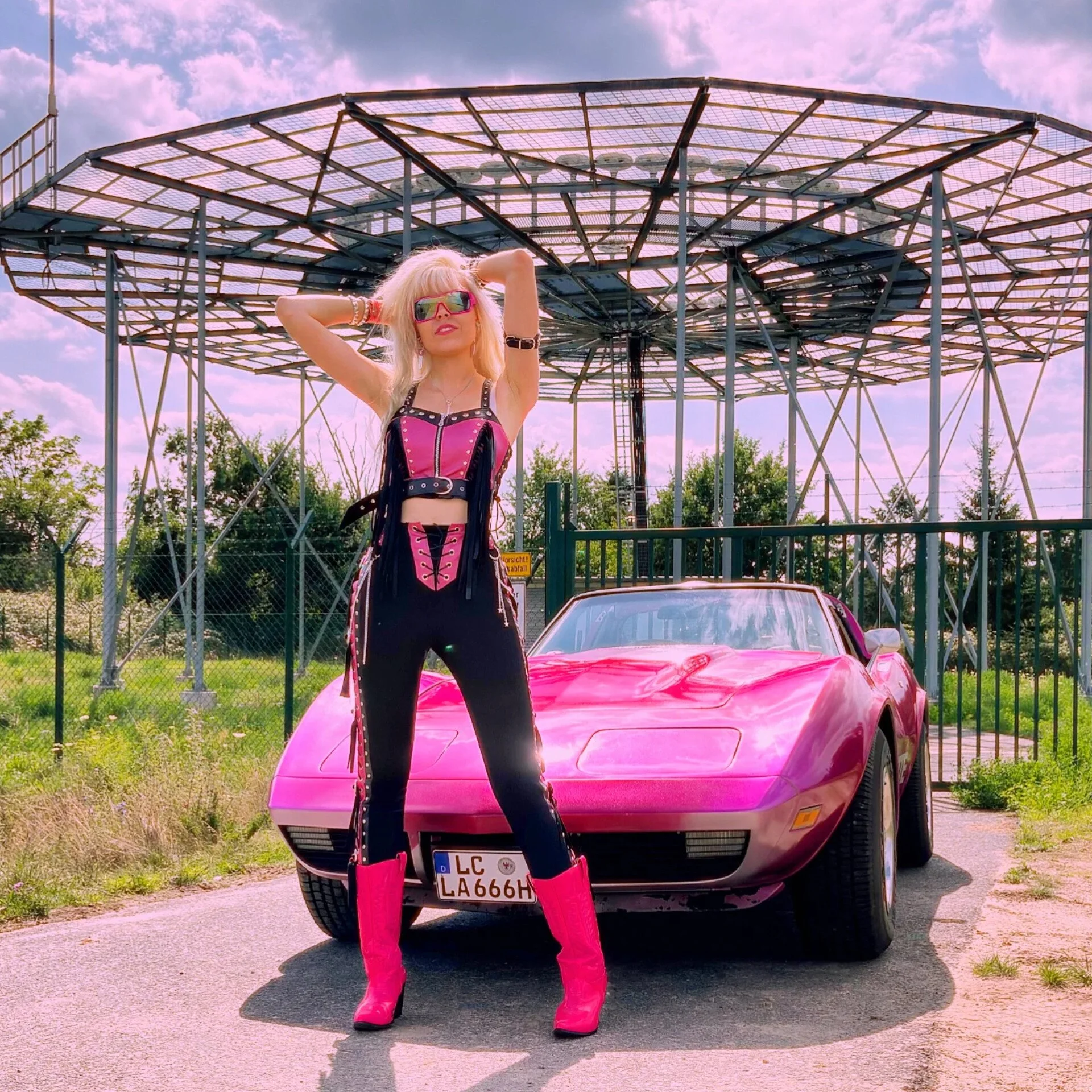 A woman with platinum blonde hair wearing a pink and black outfit with pink boots and sunglasses posing in front of a pink sports car under a large amusement park ride.