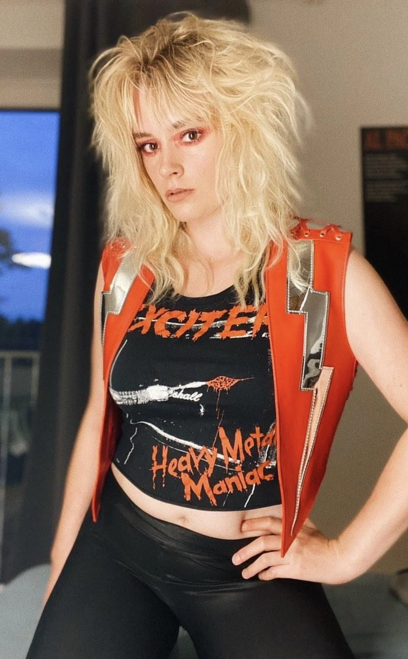 A woman with blonde, curly, messy hair wearing a black T-shirt with red text that reads 'Hated Metal Maniac' and a graphic of a guitar, paired with a red sleeveless vest with silver accents.