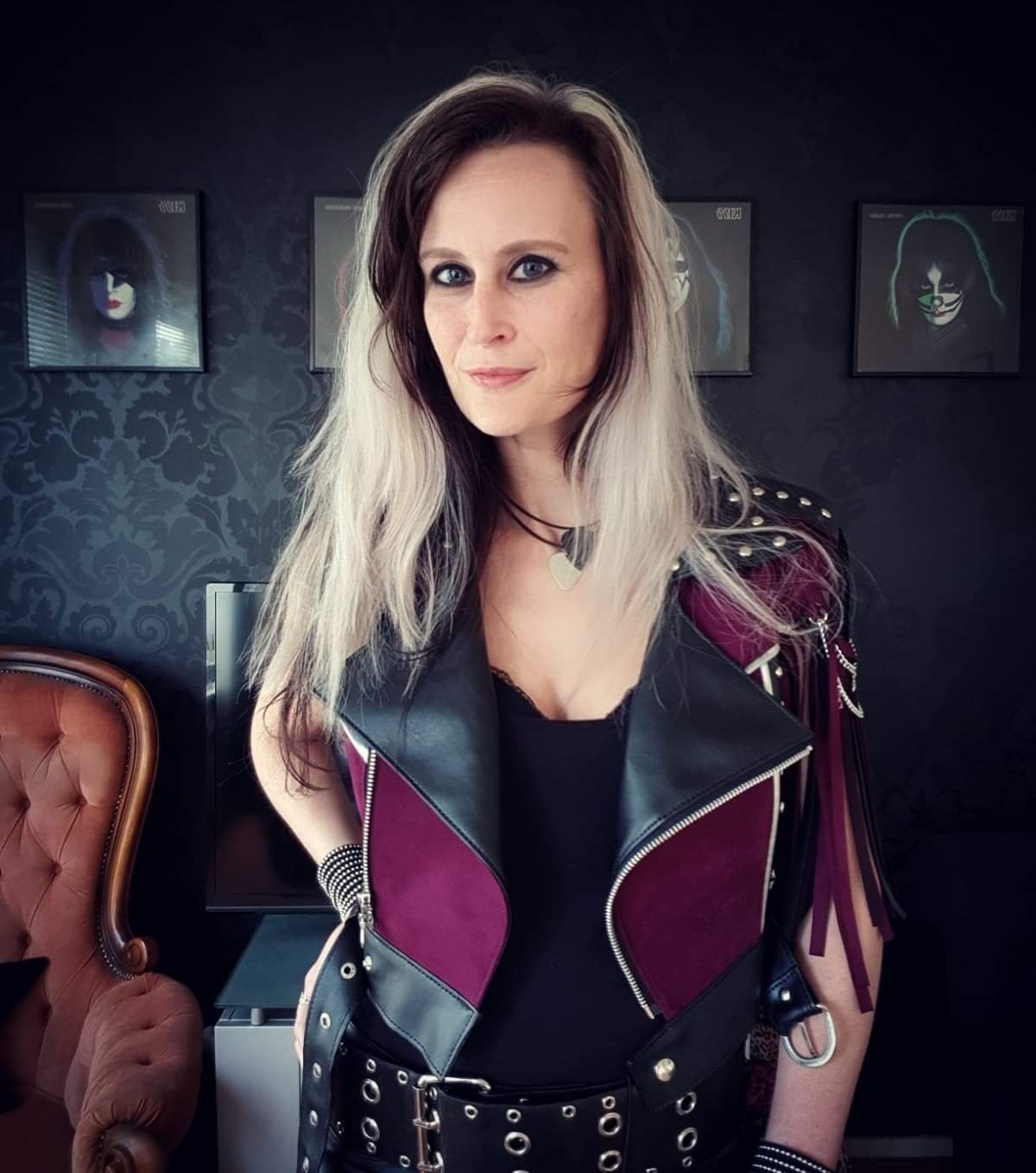 A woman with long, wavy blonde and brown hair, wearing a black top and a sleeveless leather vest with purple panels and metal studs, standing in a room with dark wallpaper and framed pictures of women with makeup and dark hair in the background.