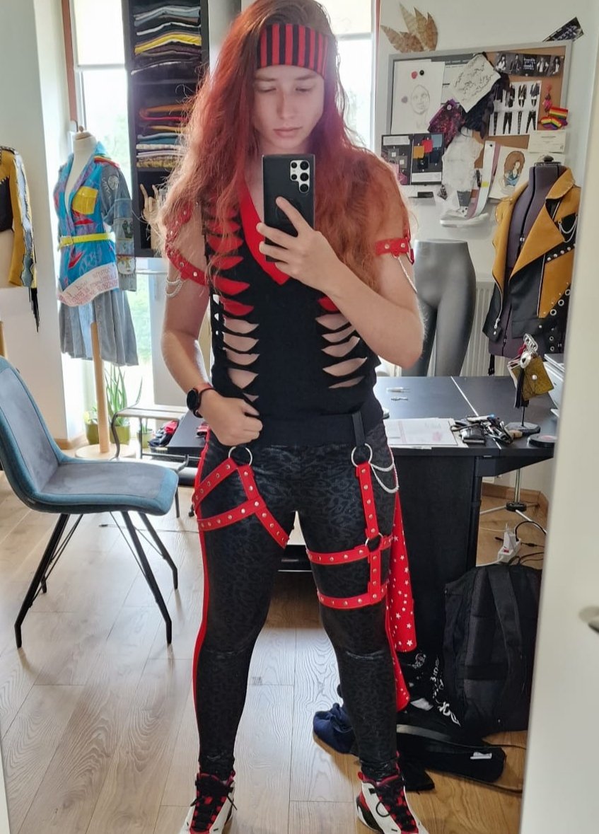A person taking a mirror selfie in a cluttered room, dressed in black and red fashion with ripped shirt and harness-style accessories, wearing a red and black headband, and sporting long reddish hair.
