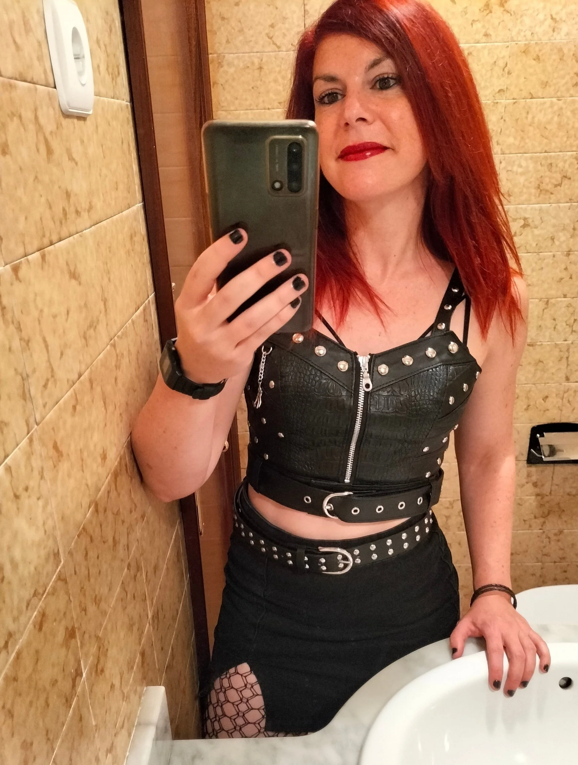 A woman with red hair taking a selfie in a bathroom mirror. She is wearing a black leather-style top with metal studs and zippers, a matching belt, a black skirt with fishnet stockings, and dark nail polish.