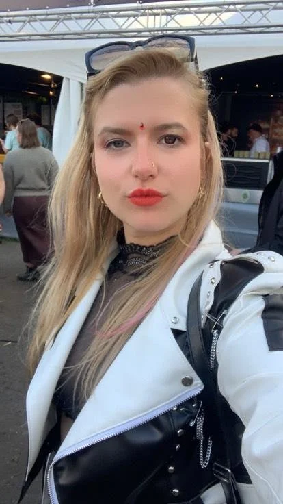 A young woman with long blonde hair taking a selfie at an outdoor event. She is wearing a white and black leather jacket, black top, gold hoop earrings, a small red bindi on her forehead, and has sunglasses on her head. There are other people and ten