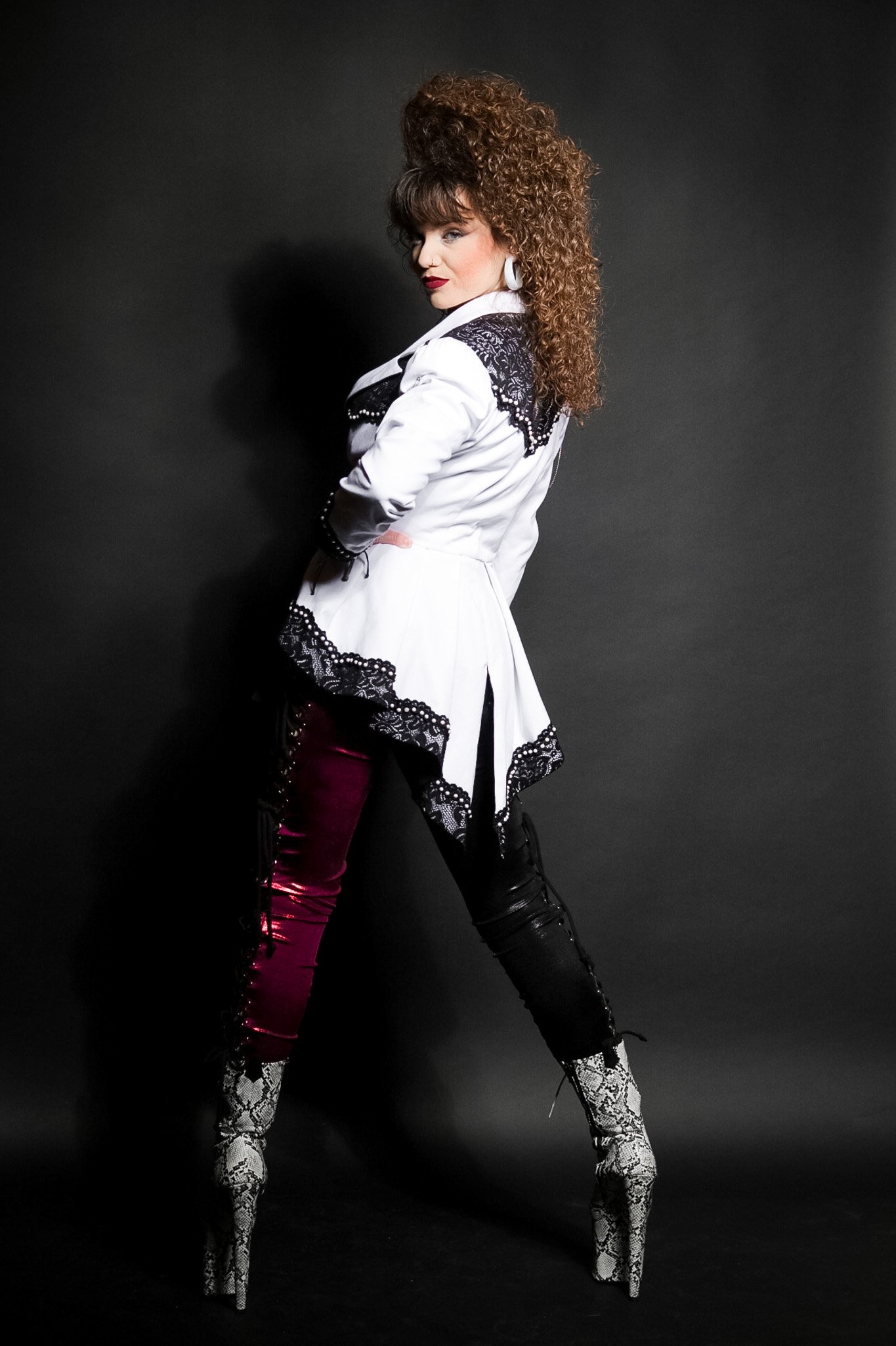 A woman with curly brown hair and red lipstick standing against a dark background, wearing a white blazer with black lace, maroon and black pants, and high-heeled snake print boots.