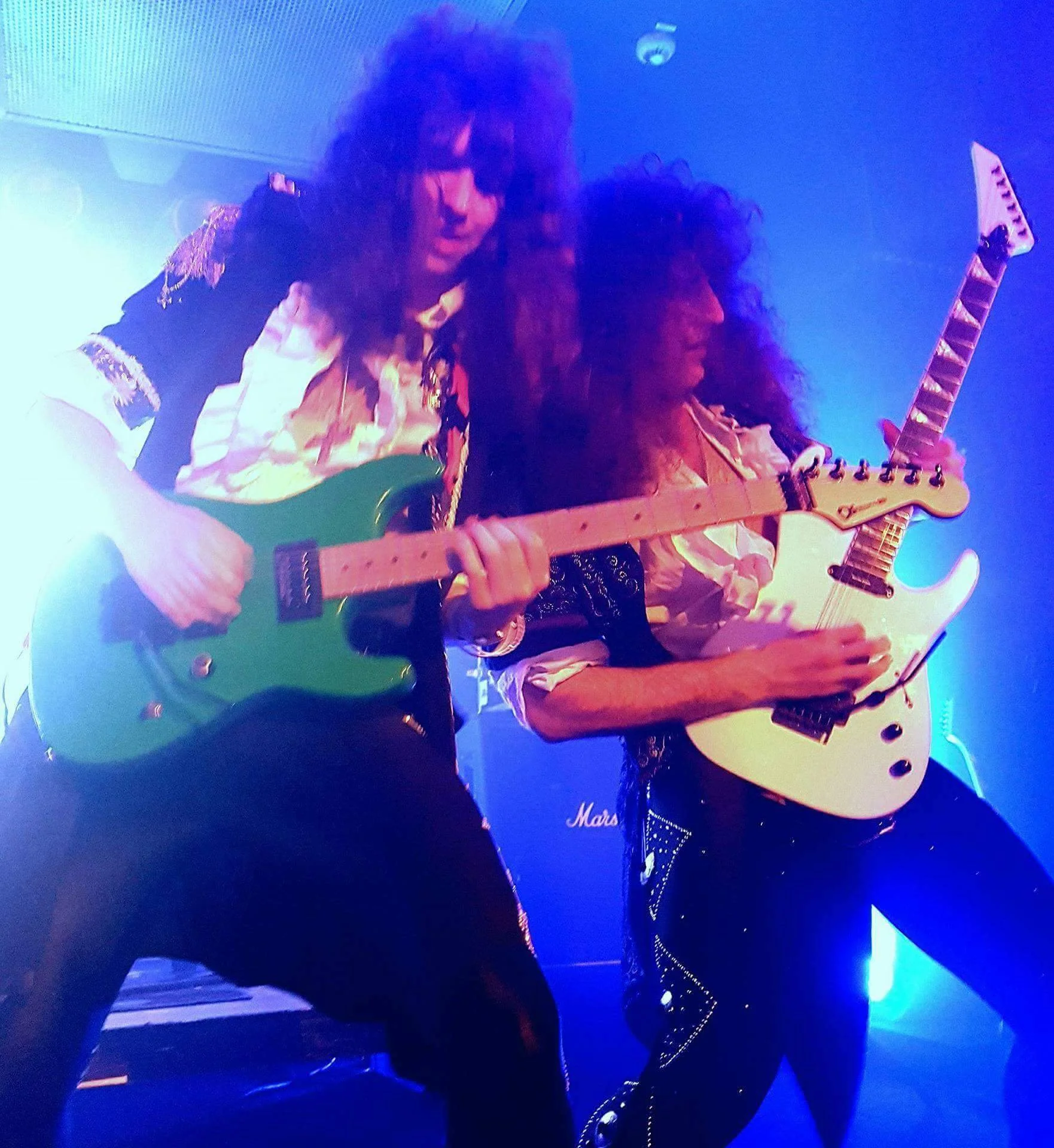 Two male guitarists performing on stage with colorful lighting, both playing electric guitars—one green and one white.