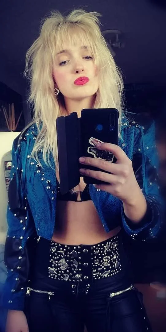 A woman with platinum blonde, messy, voluminous hair taking a mirror selfie with her phone. She is wearing a blue studded jacket, a black top with a harness, and decorated high-waisted pants with studs and embellishments.