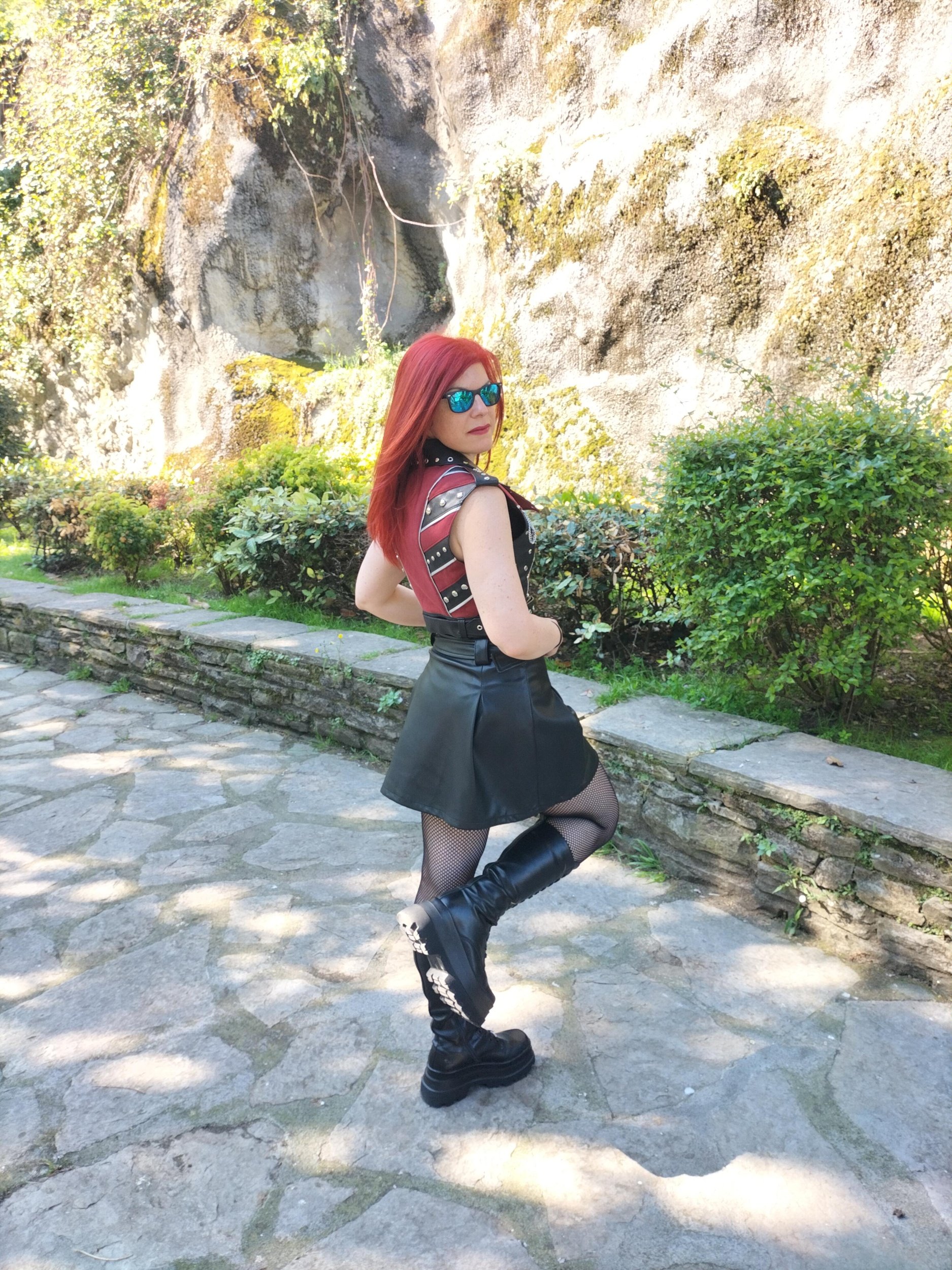 Woman with red hair and sunglasses posing outdoors on a stone pathway, wearing a sleeveless top with metal studs, a black leather skirt, fishnet stockings, and tall black boots, with a rocky wall and greenery in the background.