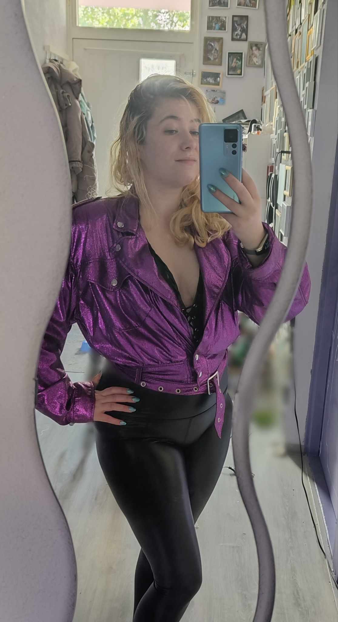 A young woman with blonde hair taking a mirror selfie, wearing a shiny purple jacket and black pants, in a room with framed photos on the wall.