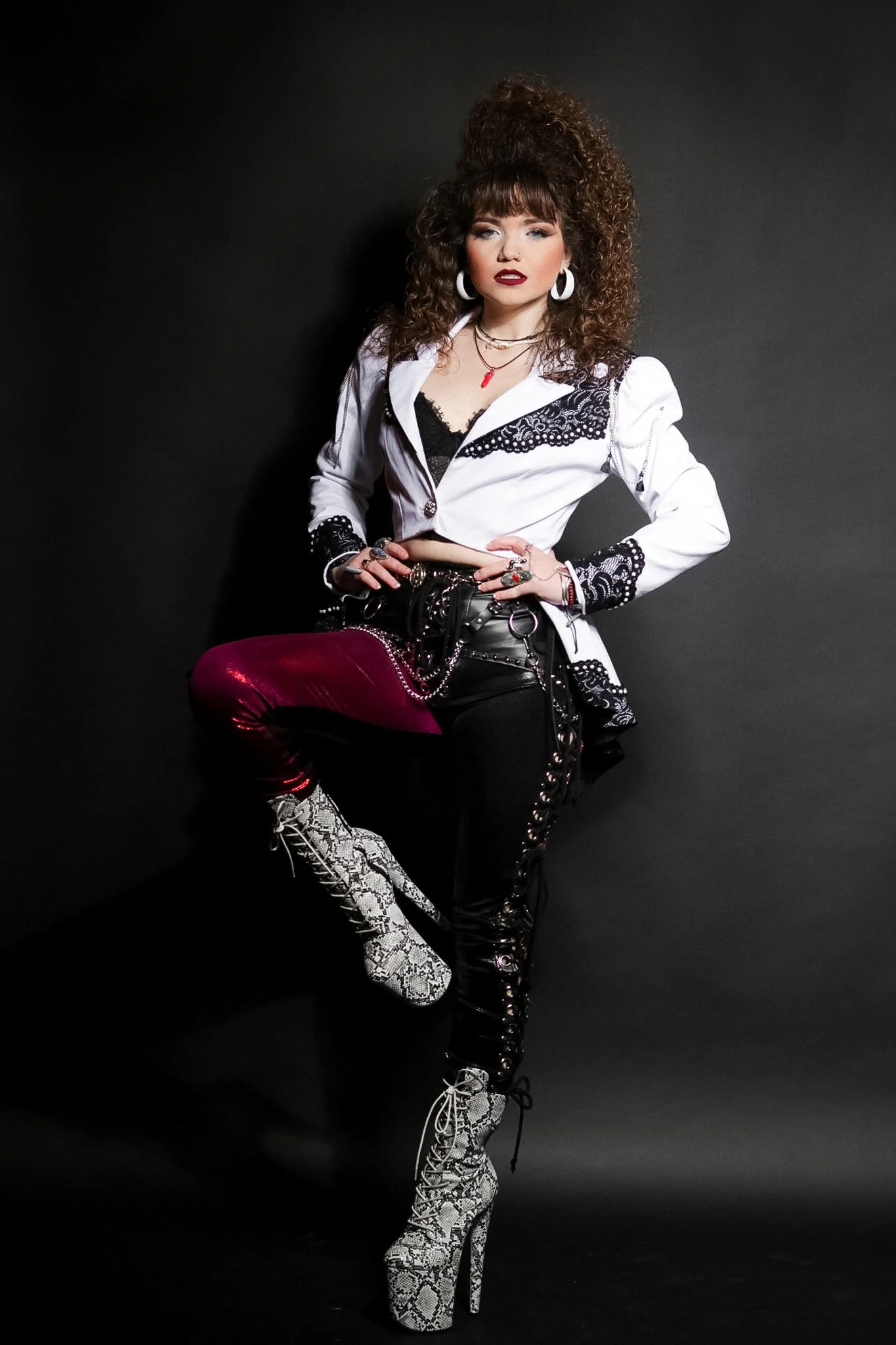 A woman with curly hair wearing a white and black lace jacket, black pants with lace-up details, snakeskin high heel boots, and bold accessories poses against a black background.