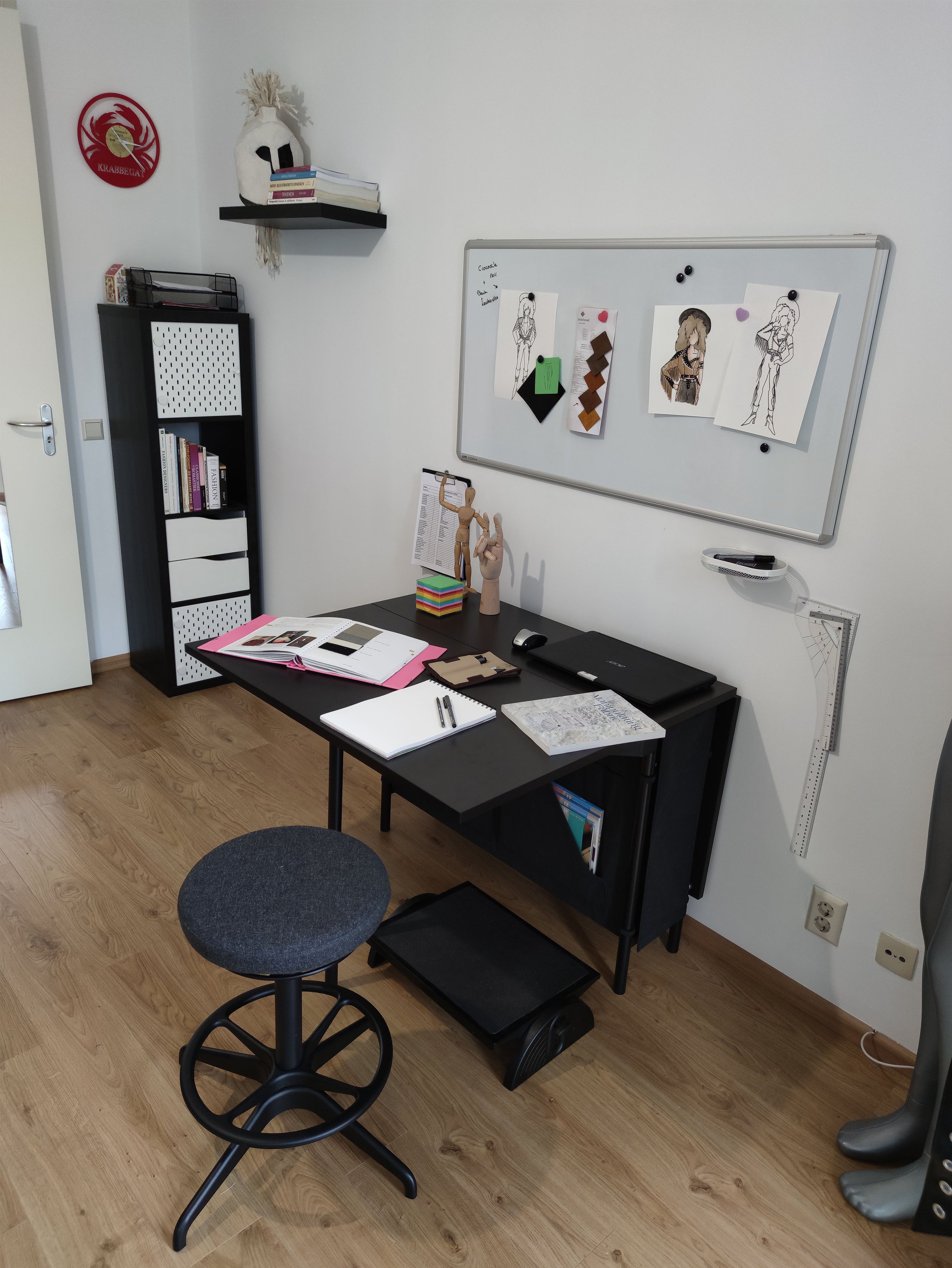 A modern home office with a black desk, a gray stool, and a whiteboard on the wall. The desk has books, notebooks, and a laptop. There is a black bookshelf with books and baskets, and decorative items including a wooden mannequin and paper models. The wall has posters, samples, and small decorations, with a feathered white mask on a shelf.