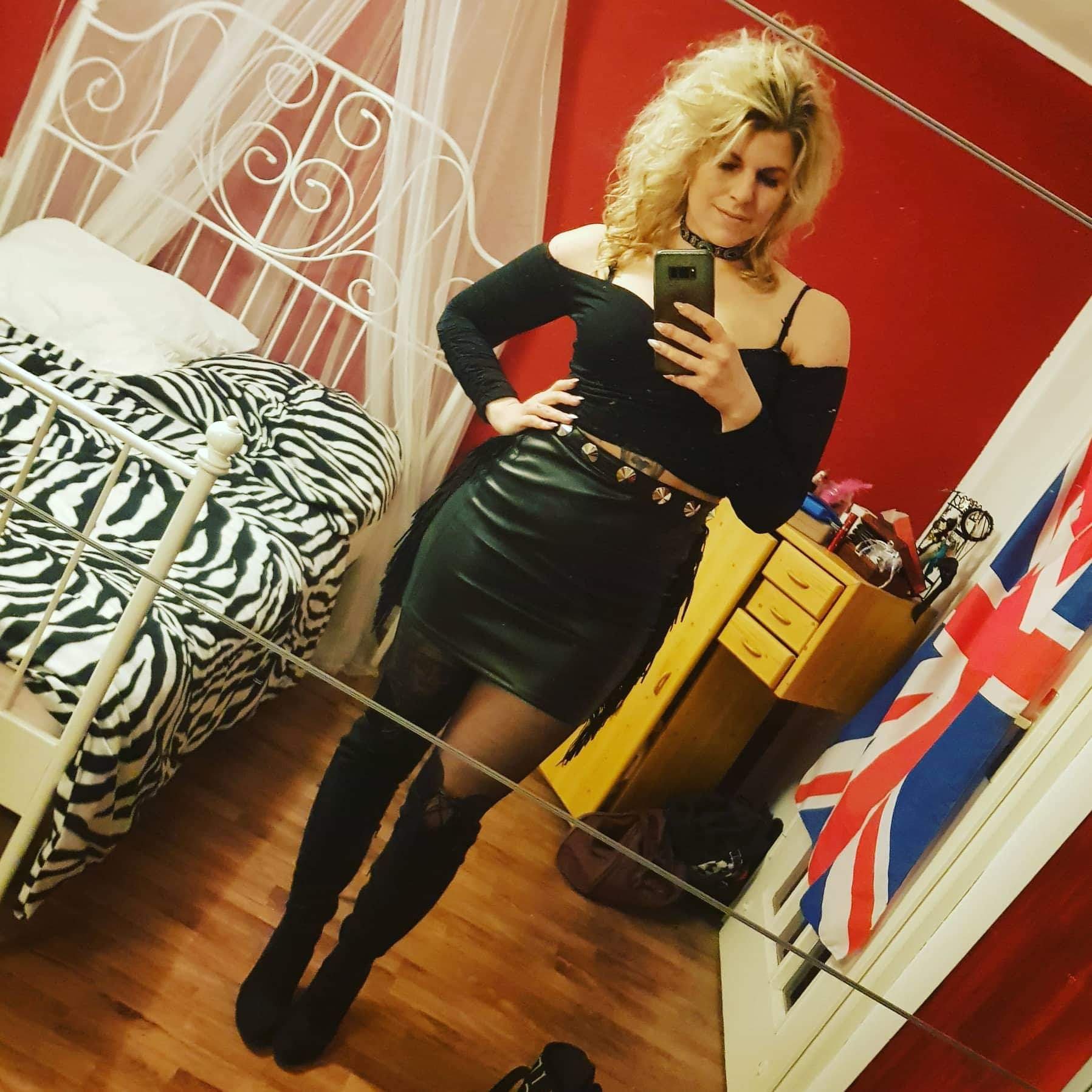A woman taking a selfie in a bedroom with red walls, a white metal bed frame with a zebra print blanket, a wooden dresser, and a British flag hanging on the wall.